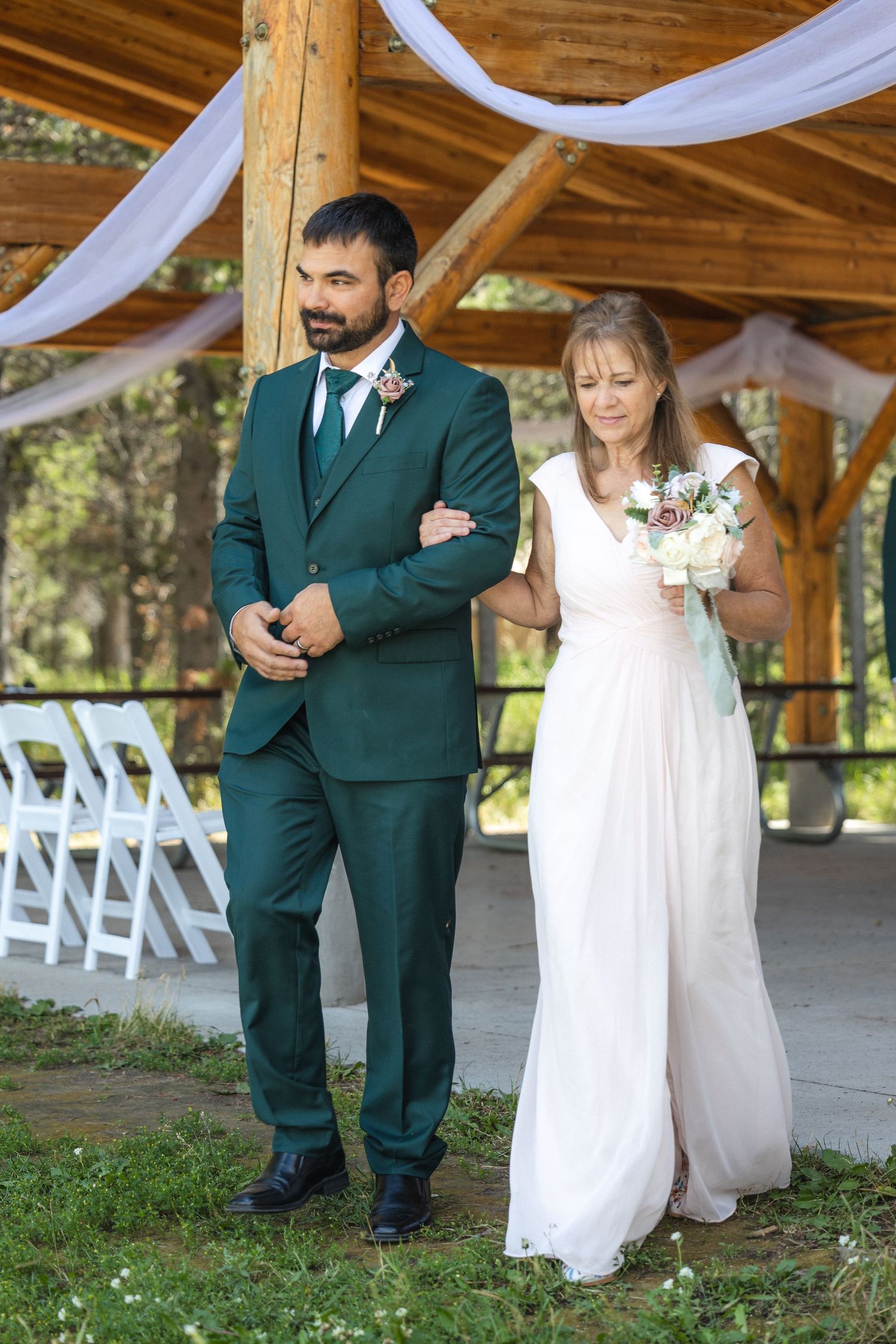 Couple in wedding attire standing under rustic wooden pavilion decorated with white draping.