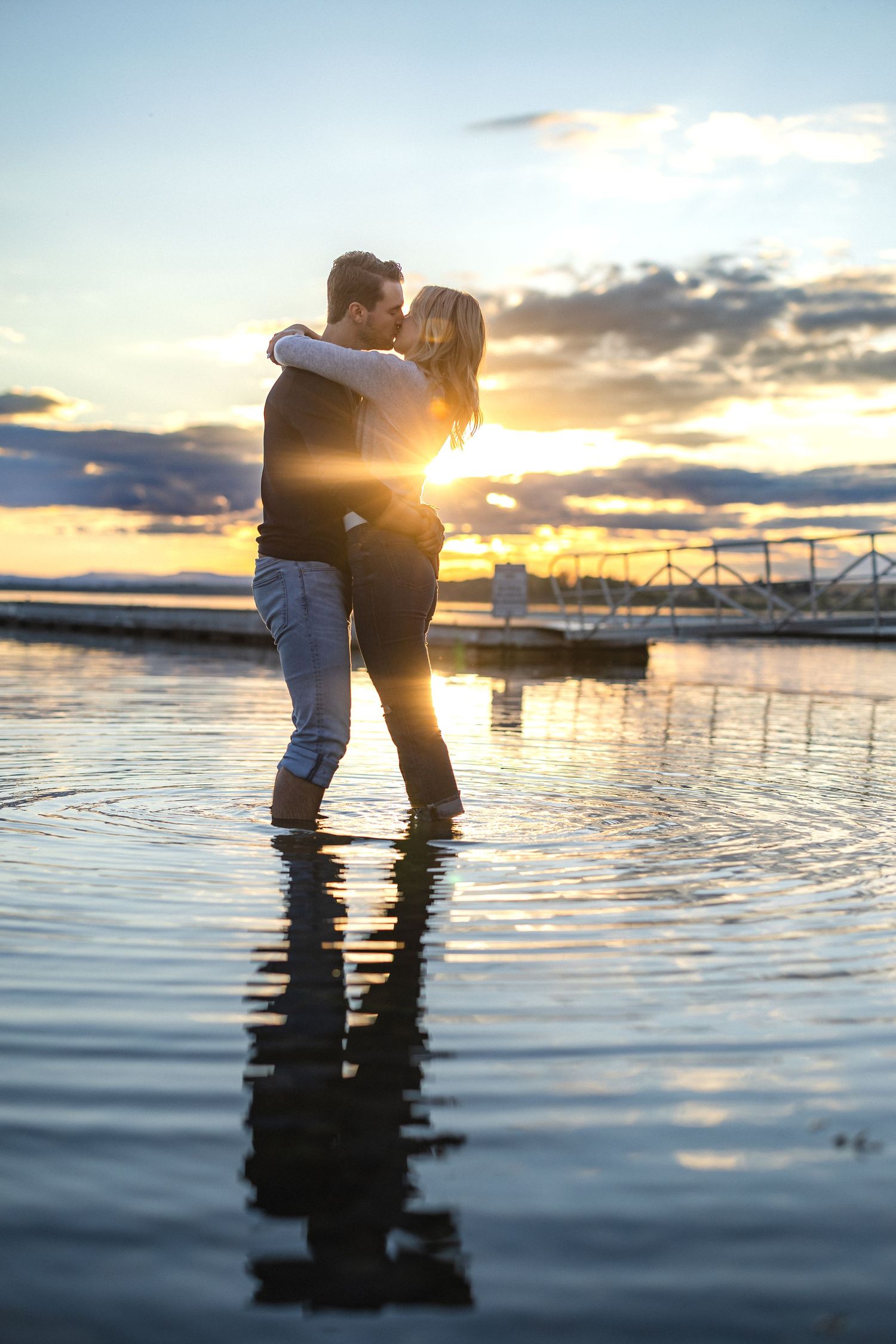 Silhouetted couple kissing in water with golden sunset and marina backdrop.