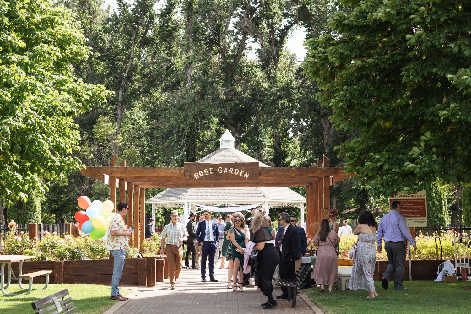 Wedding guests gather outside wooden archway and gazebo in garden setting.
