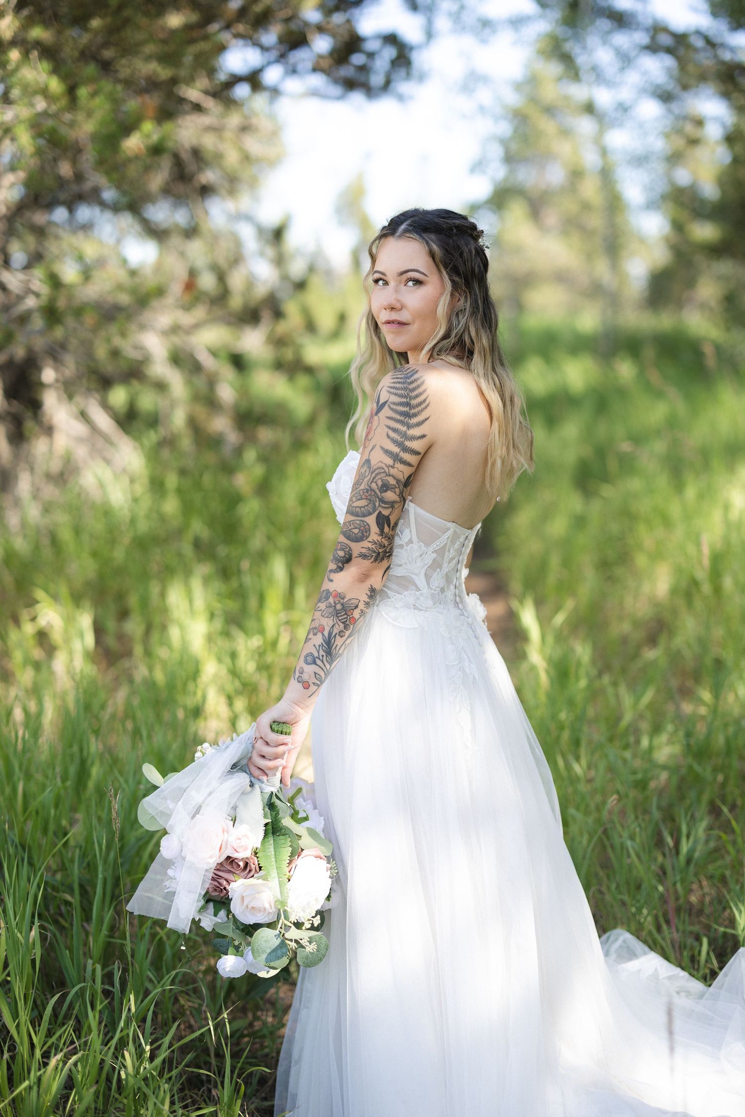 Bride in white lace gown poses gracefully in a sunlit forest meadow.