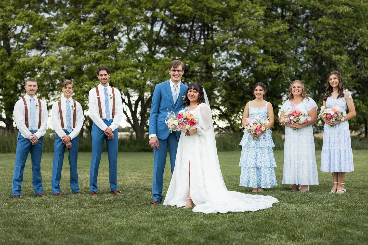 Bridal party in coordinated blue and white attire posing outdoors on grass.