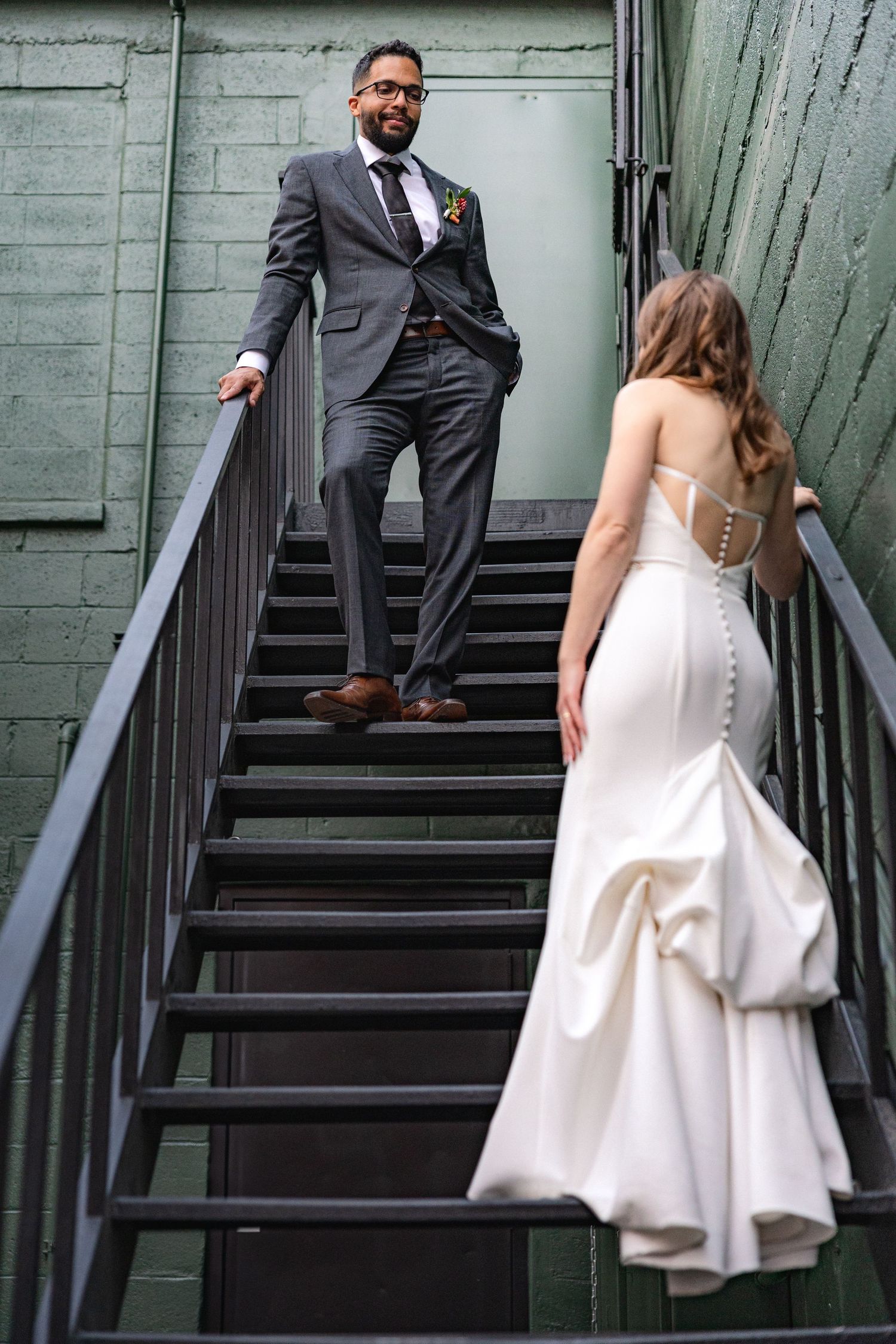 A couple poses dramatically on an industrial metal staircase with a green wall backdrop on their wedding day.