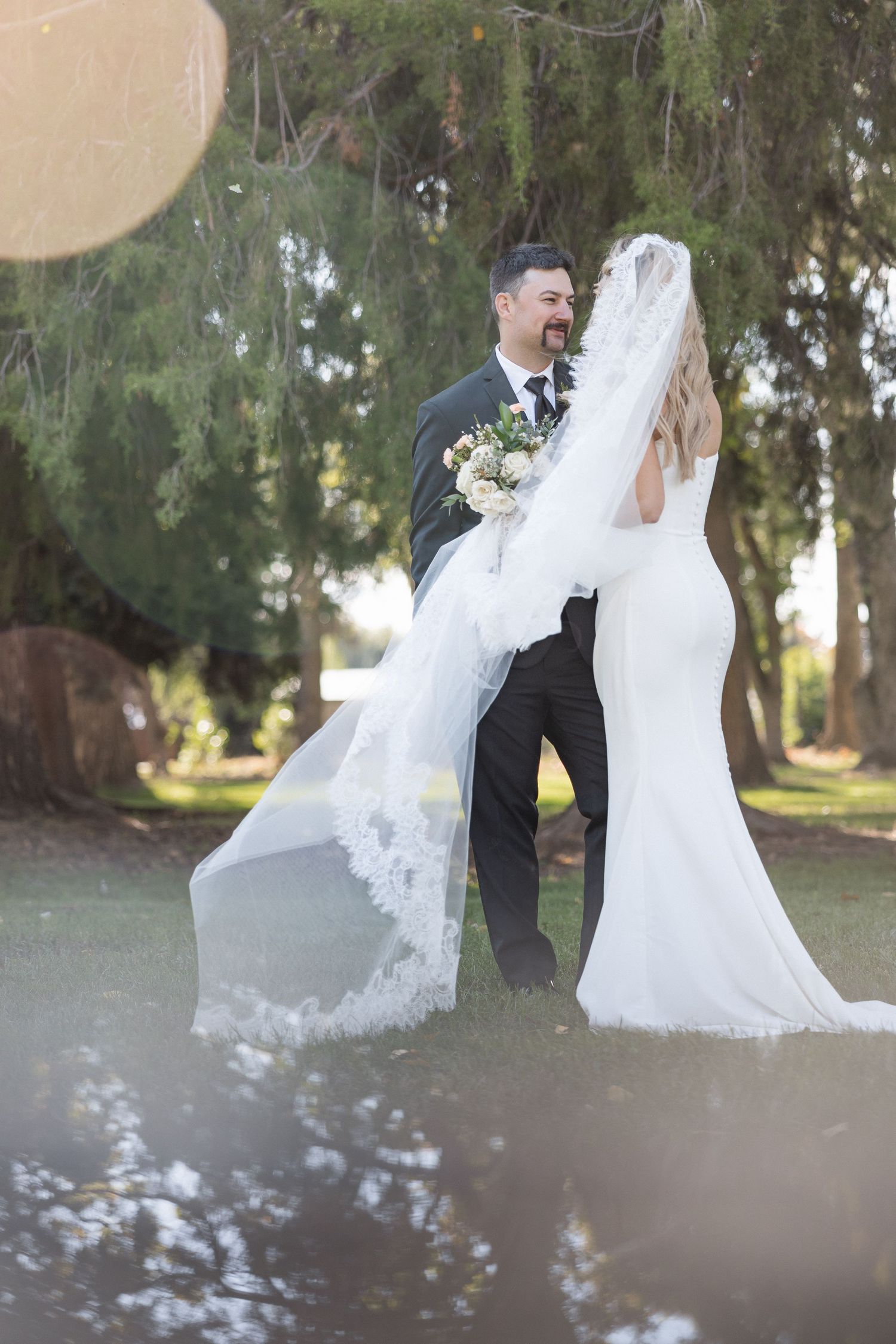 A flowing white wedding veil catches the sunlight in an outdoor garden setting.