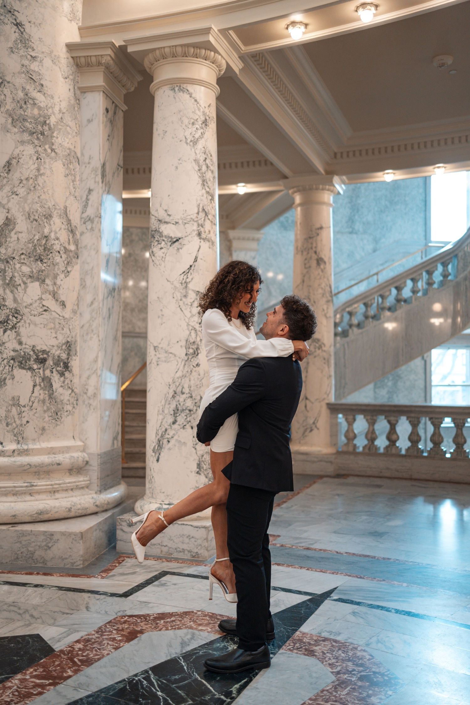 Couple embraces by marble columns in elegant building with dramatic lighting.