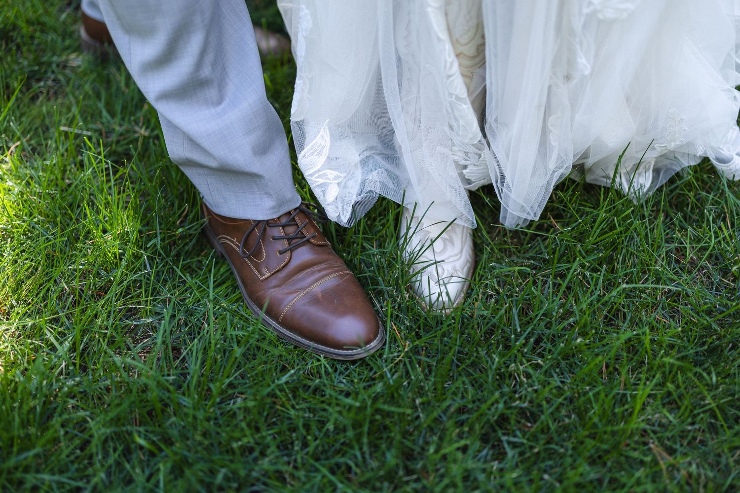 Brown leather dress shoe next to white wedding dress hem on grass.