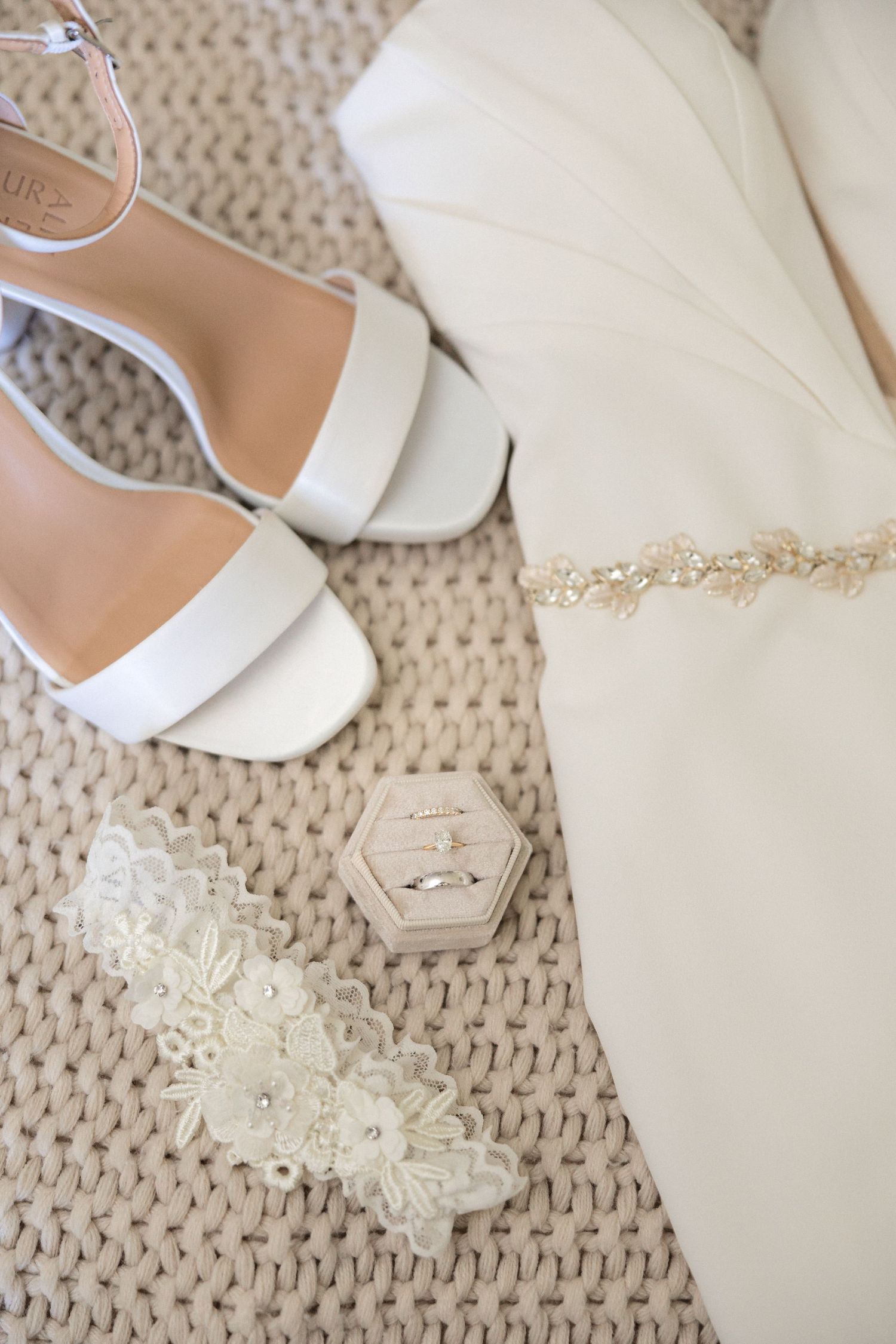 Wedding accessories including white heels and lace garter arranged on textured neutral fabric.