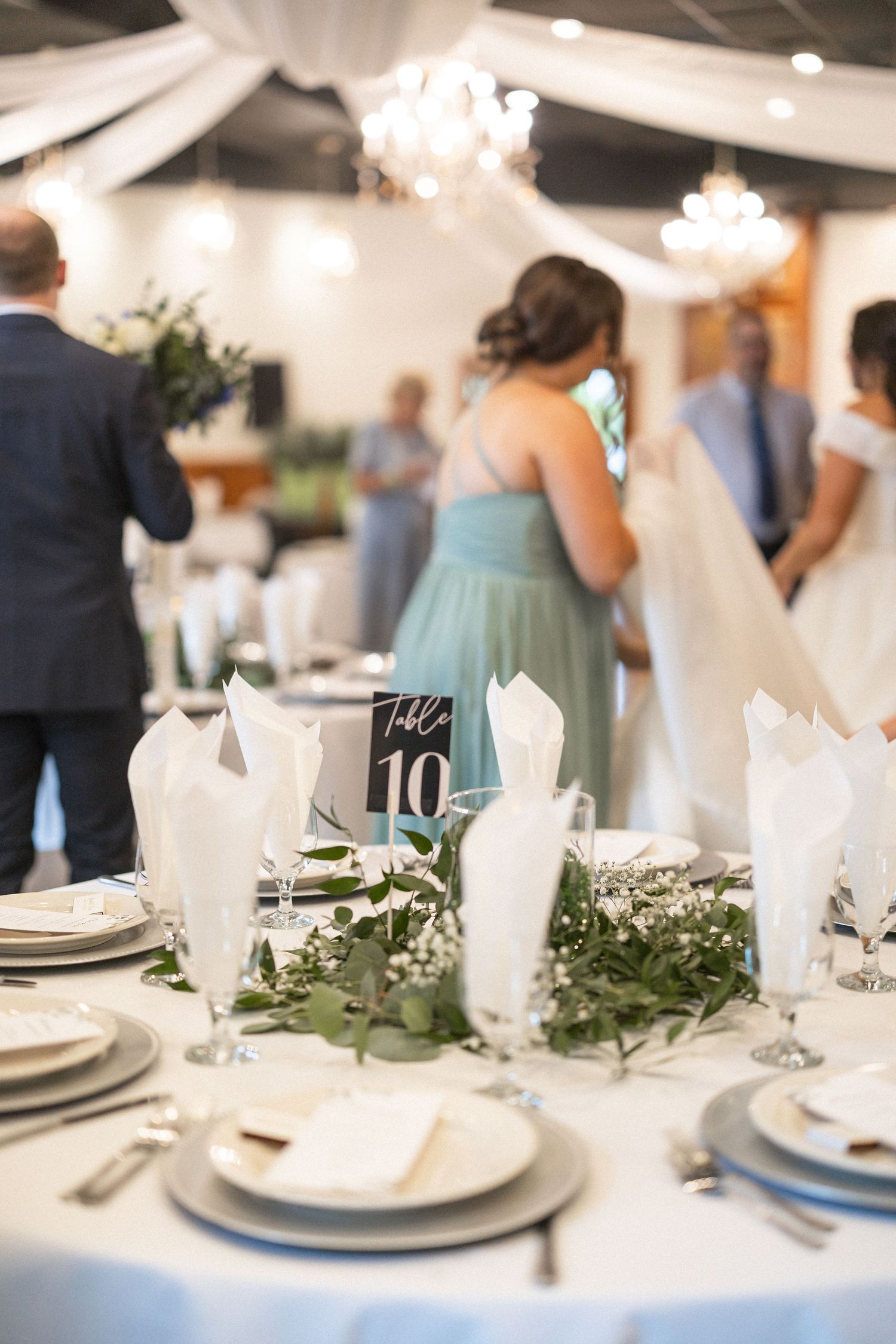 An elegant wedding reception table setting with white napkins and greenery centerpieces.