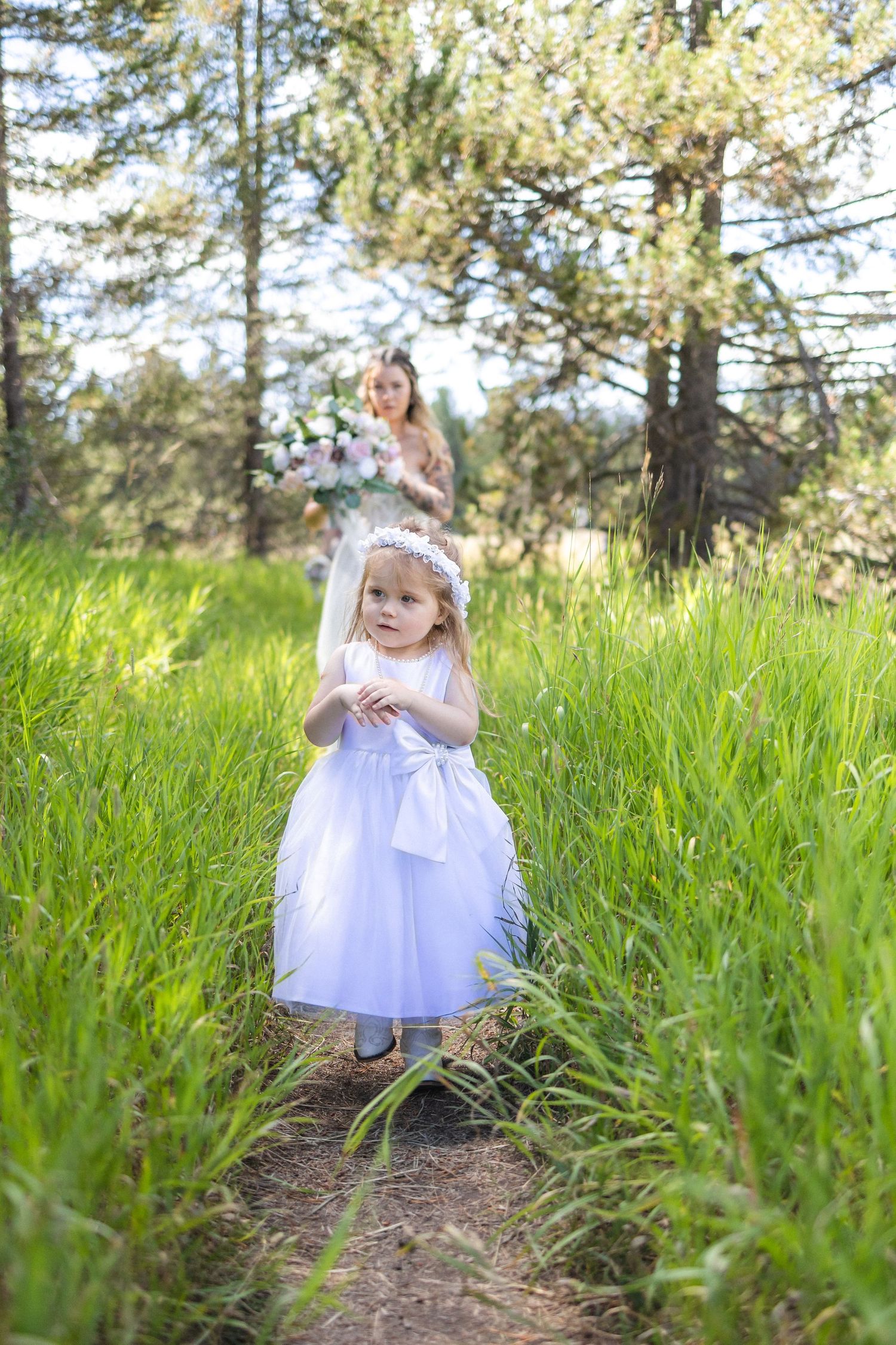 Little flower girl in white dress walks path through tall grass with wedding backdrop.
