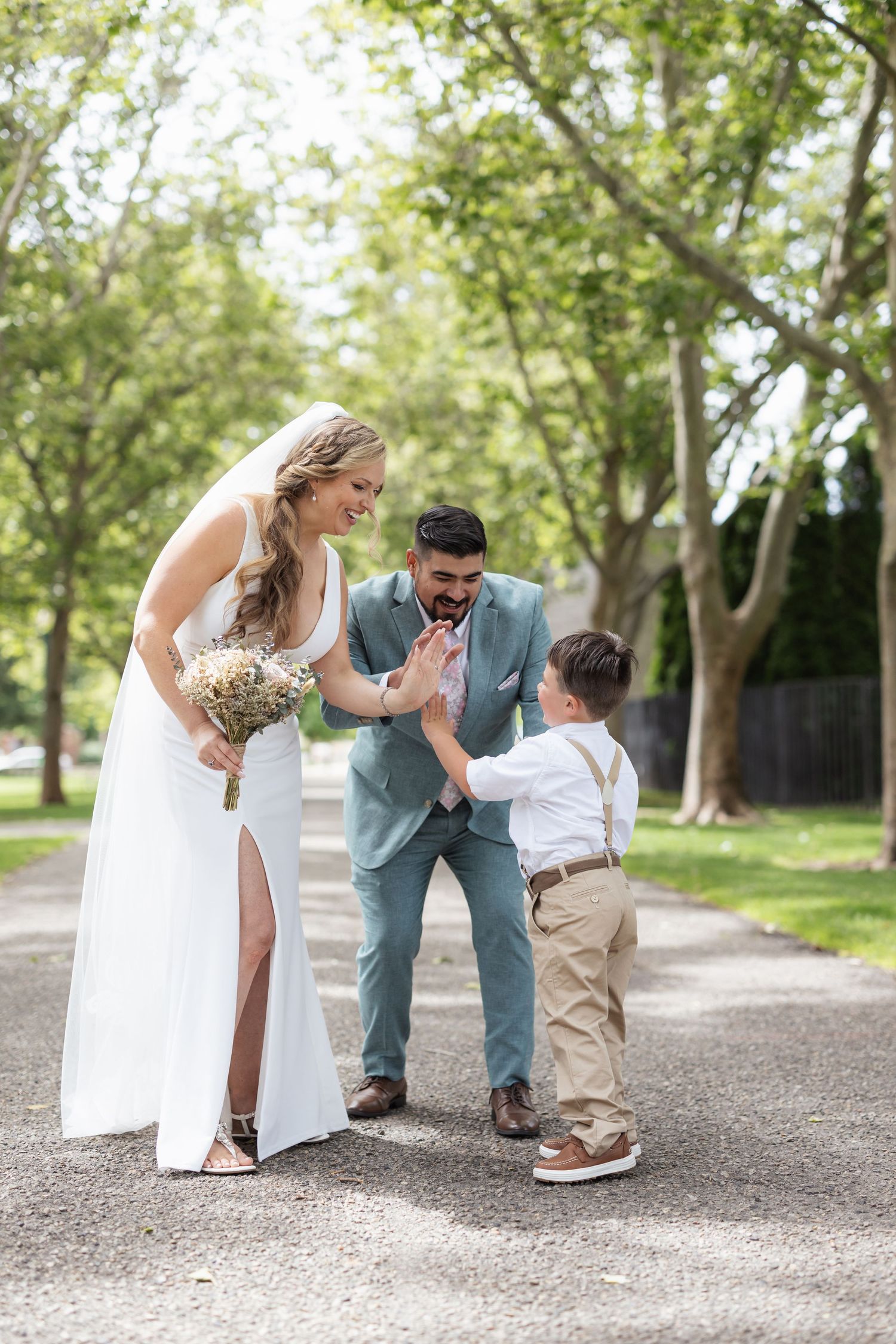 Newlyweds in white dress and light blue suit share a playful moment with young boy on tree-lined pathway.