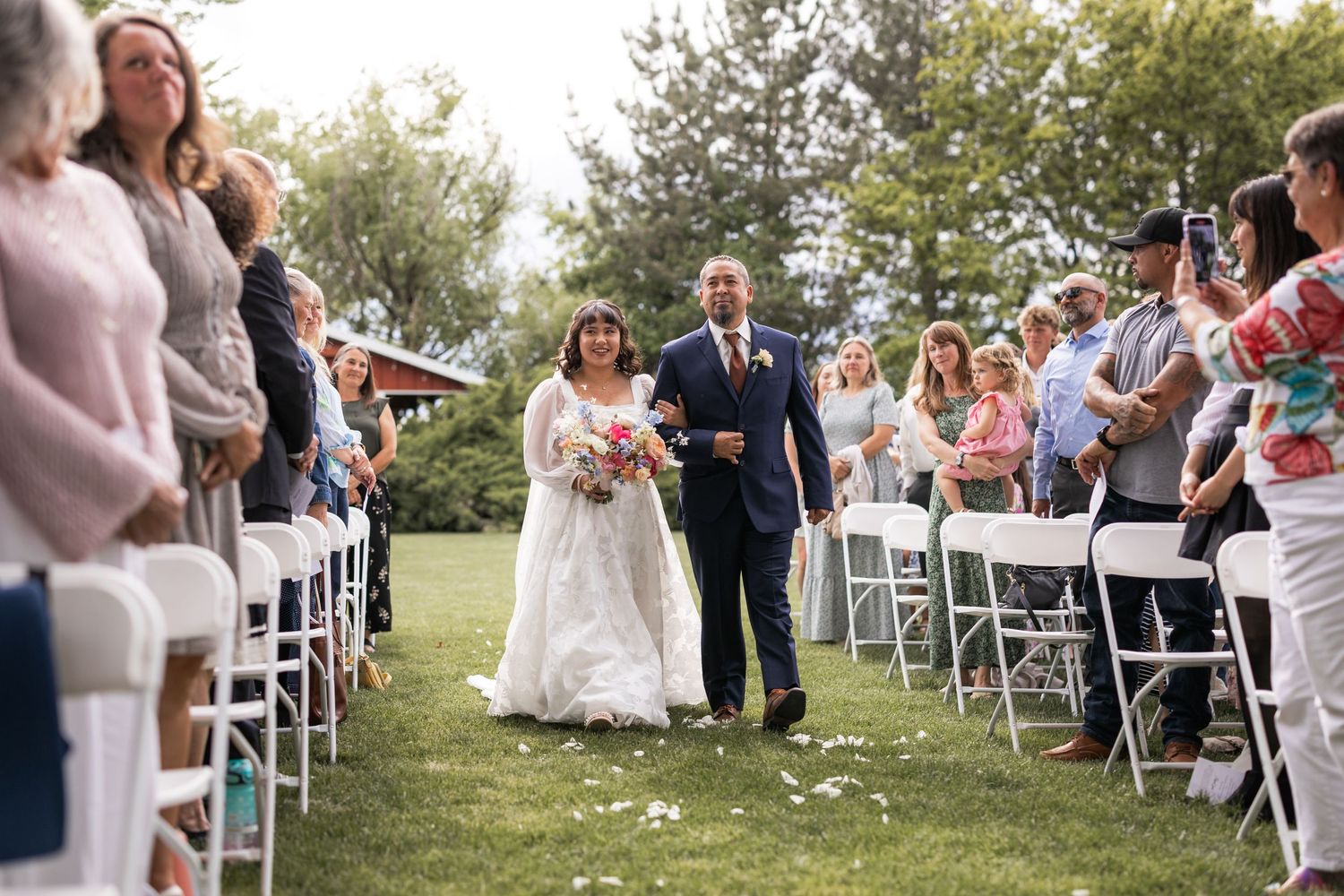 Bride walking down outdoor aisle with escort as wedding guests look on from white chairs during garden ceremony.