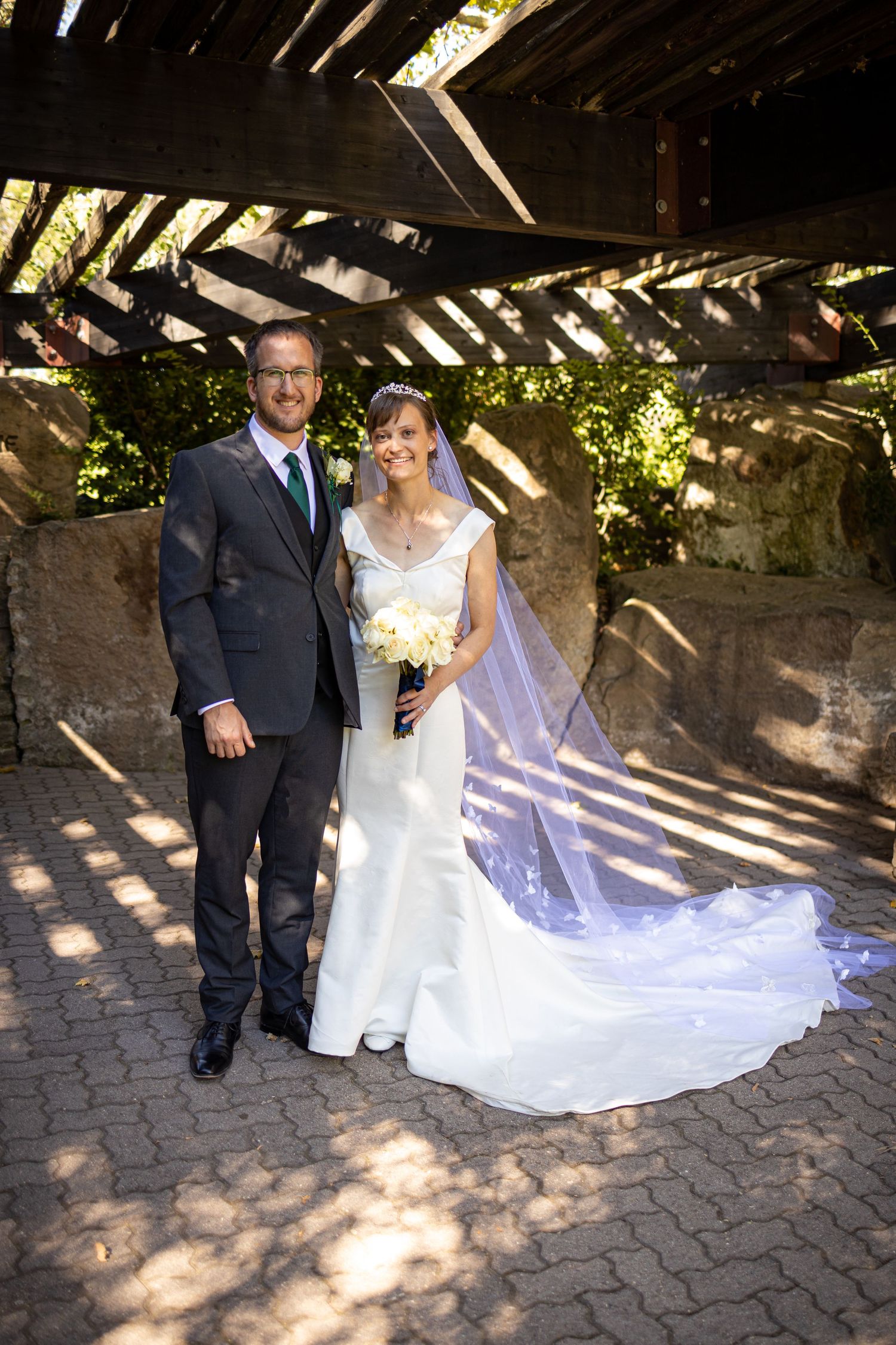 Wedding couple poses for a portrait under a wooden pergola with dappled sunlight.
