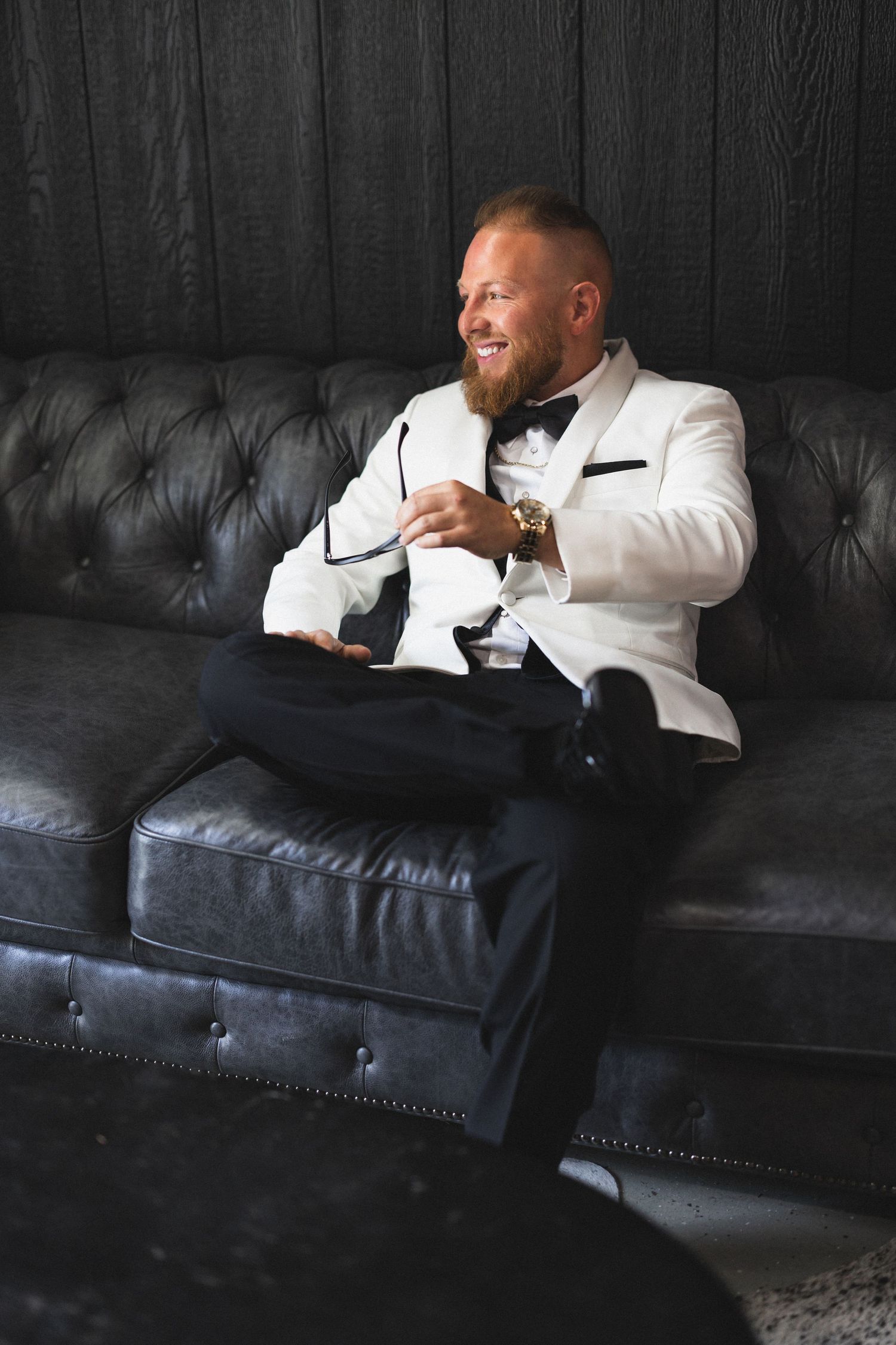 Groom relaxes on dark leather couch wearing white dinner jacket.