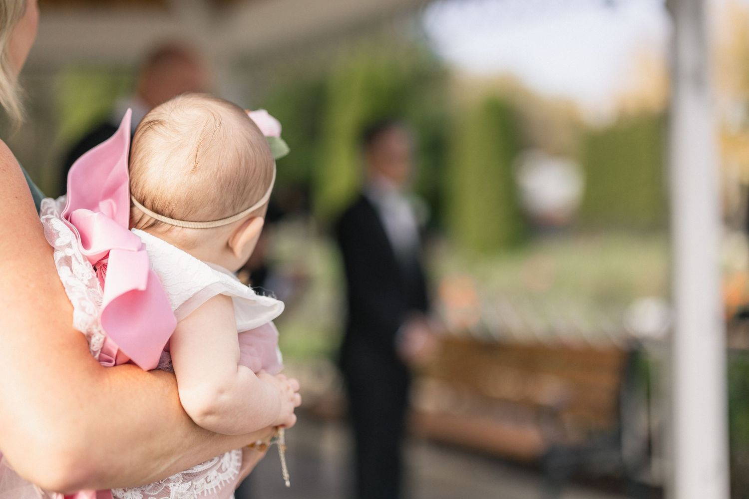 Baby in pink dress being held during outdoor wedding ceremony.