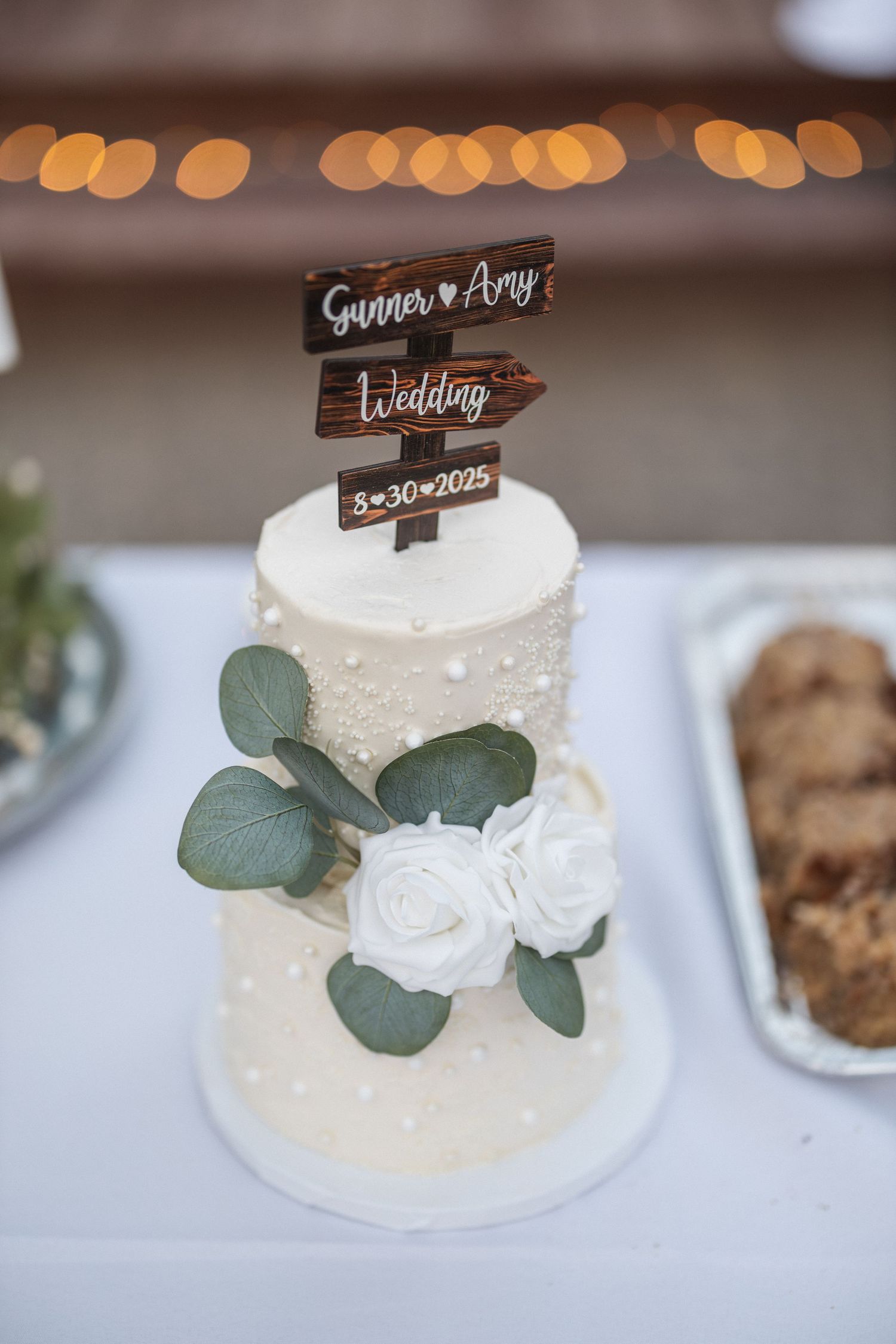Elegant two-tier white wedding cake decorated with eucalyptus leaves, white roses and a rustic wooden directional sign topper.