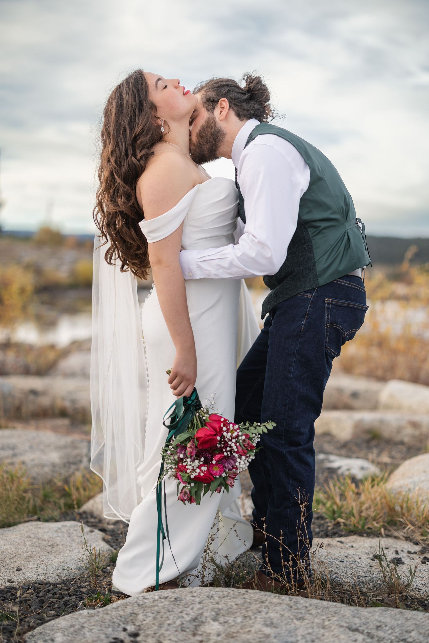 A romantic wedding couple shares a passionate kiss on rocky terrain with a beautiful cloudy sky backdrop.