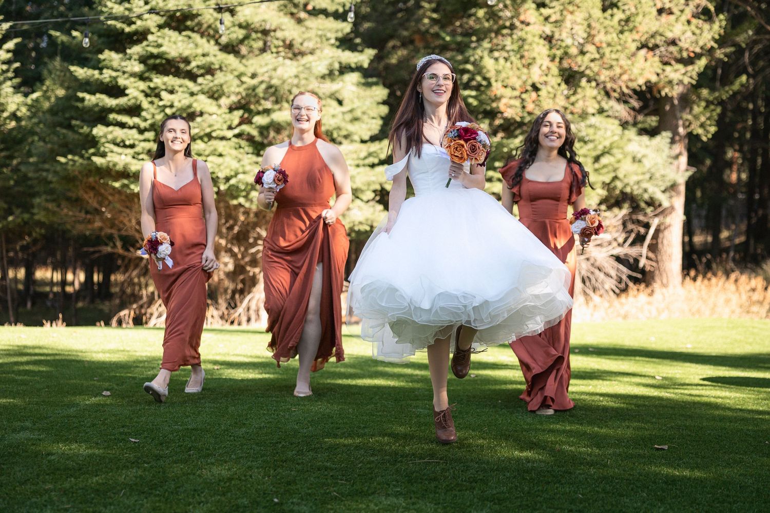 Bridesmaids in rust-colored dresses run playfully on grass with bride in tea-length wedding dress.