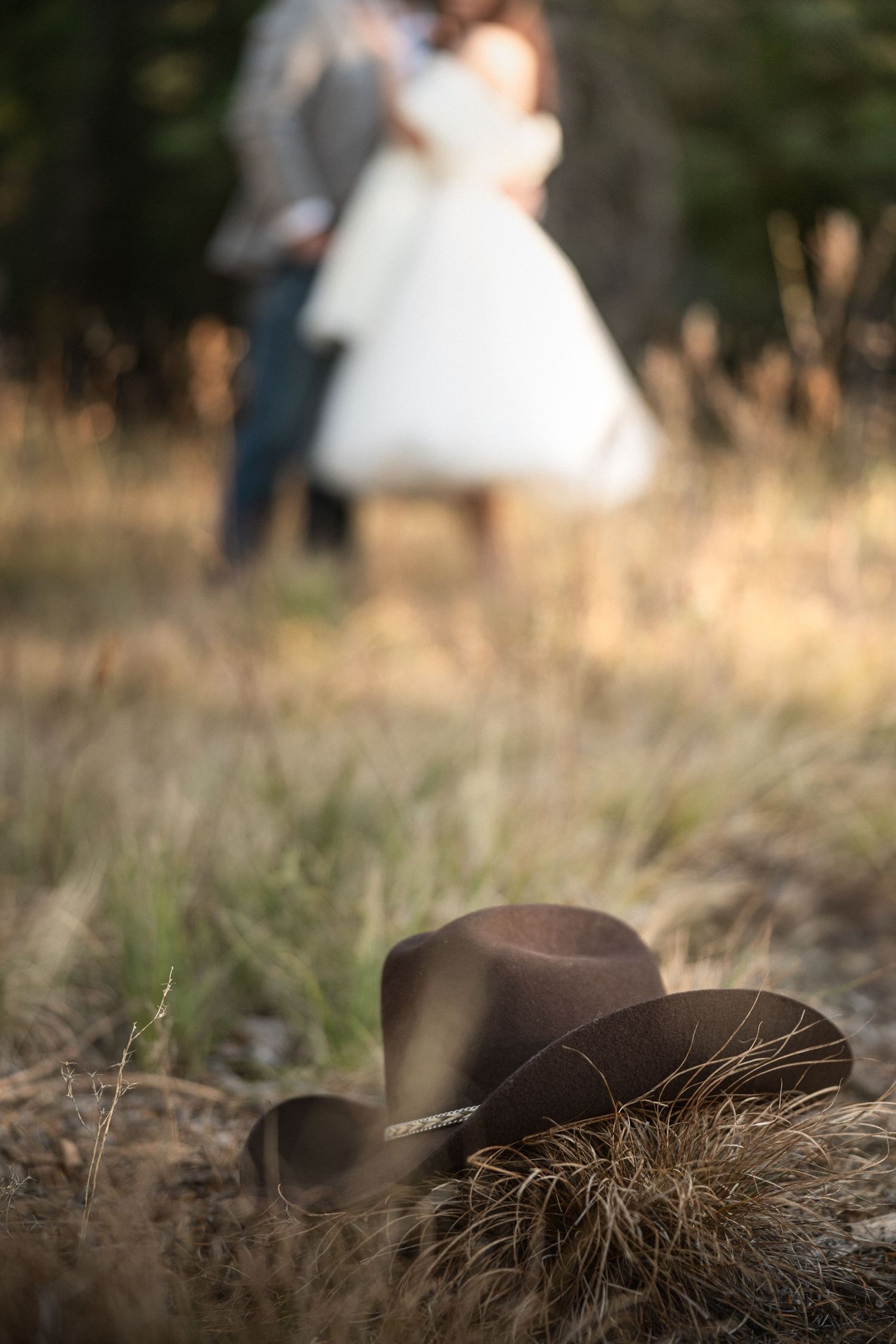 A brown cowboy hat rests on dry grass with a blurred couple in wedding attire embracing in the background.