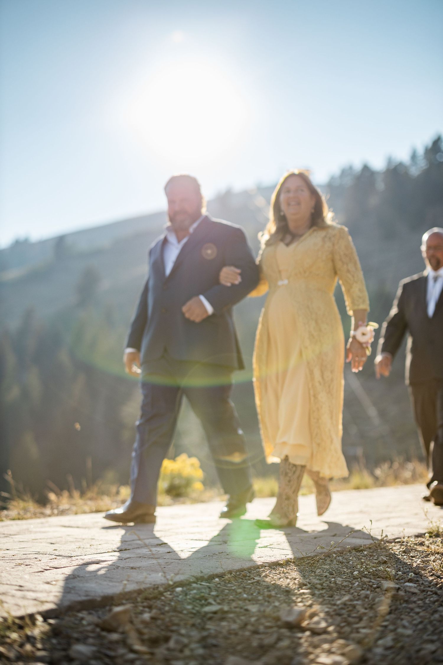 Couple walks arm in arm down a sunlit mountain path during a wedding ceremony.