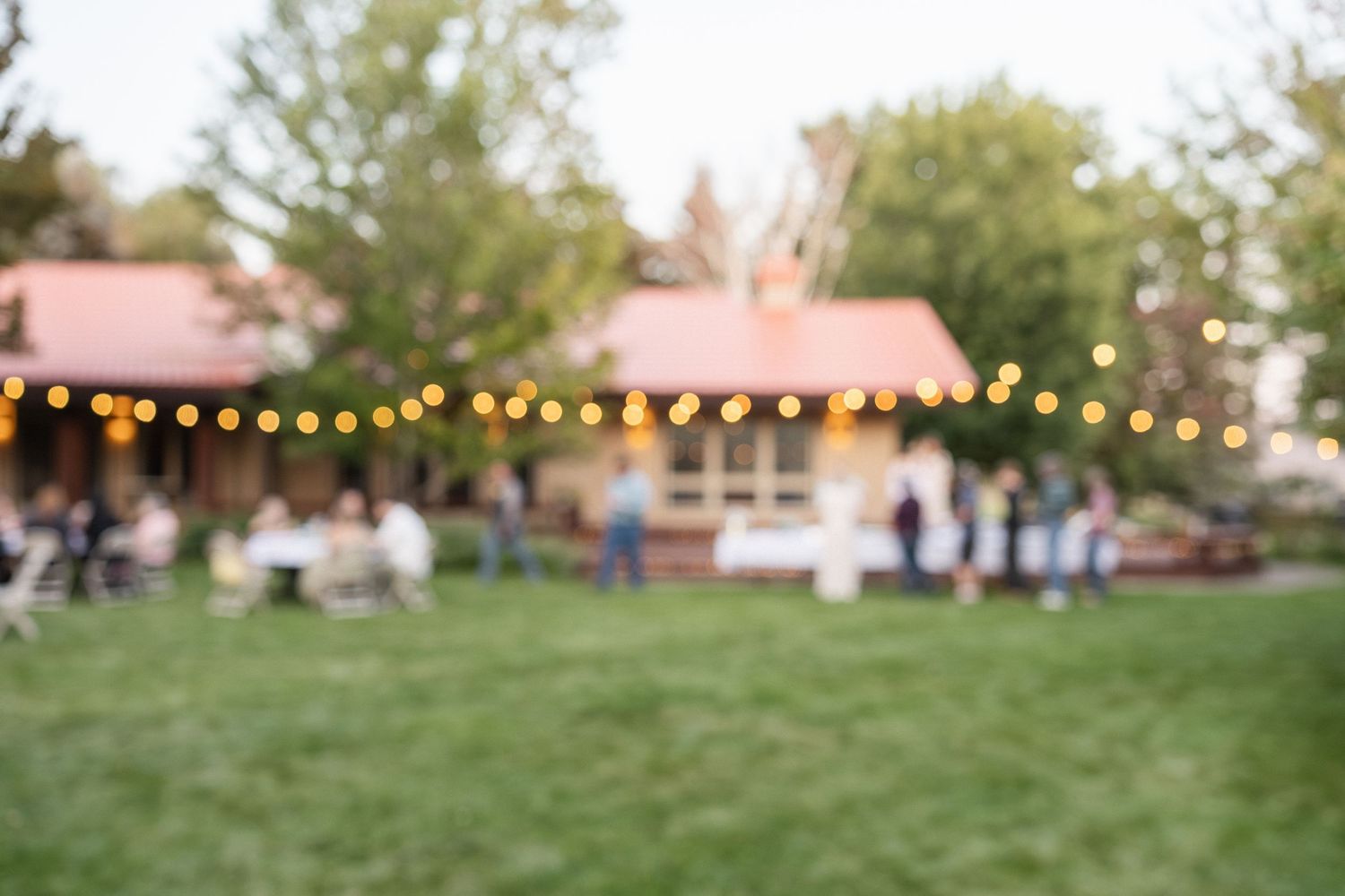 Blurred outdoor wedding reception venue with string lights and red-roofed buildings at dusk.