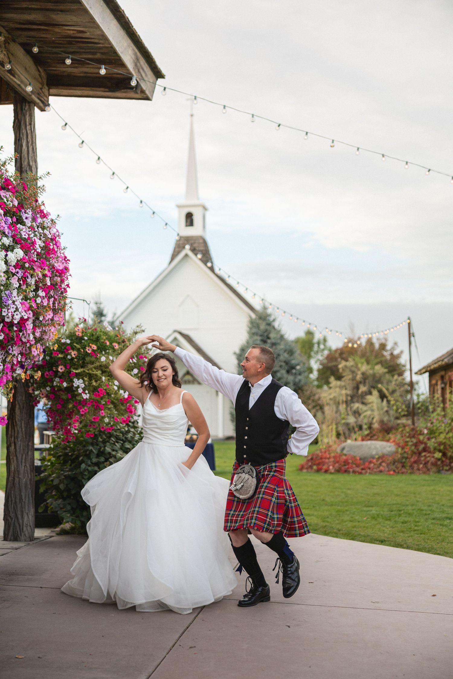 A couple dances joyfully under string lights with a white chapel and pink flowers in the background.