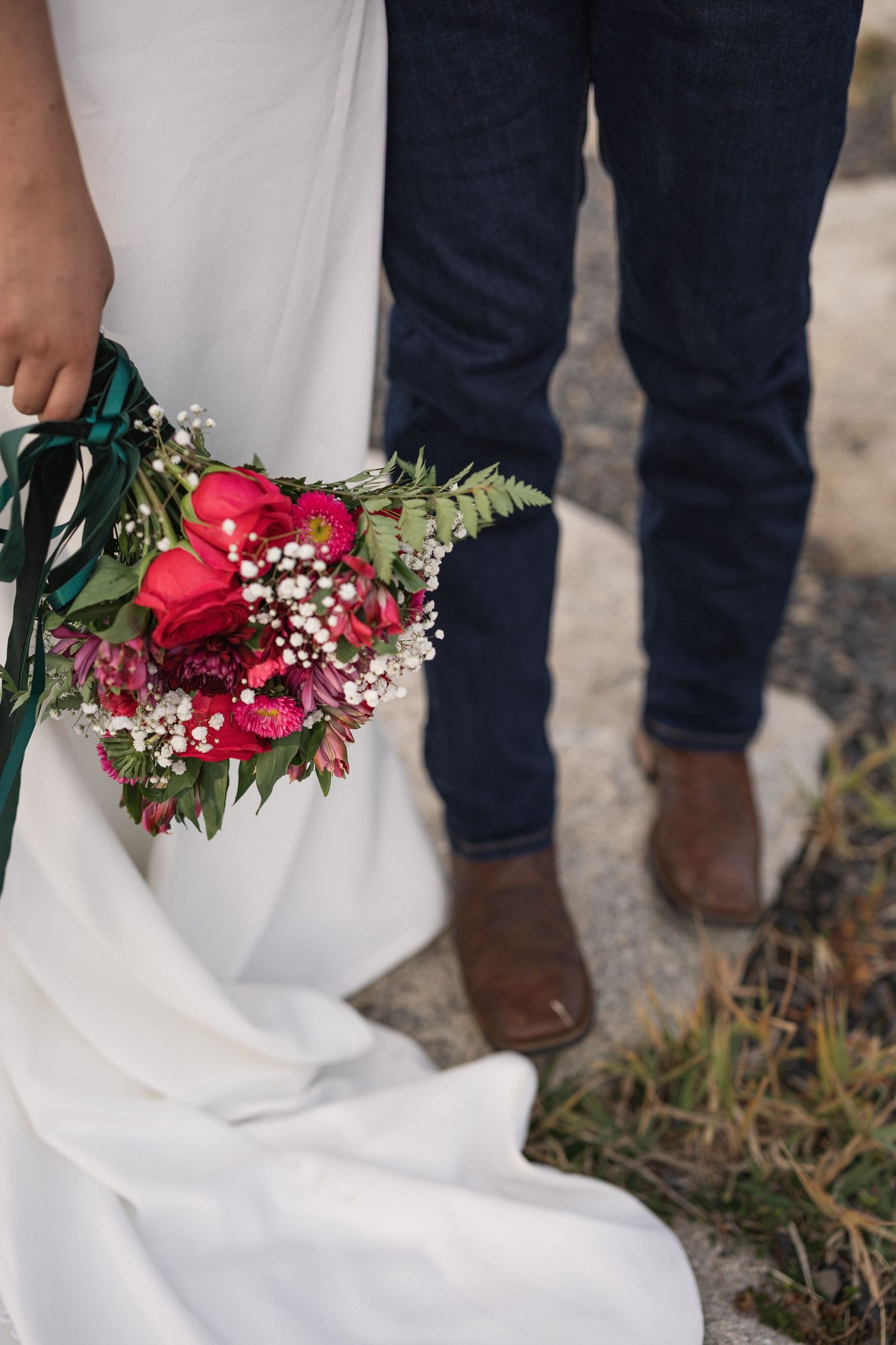 Detail shot of vibrant red wedding bouquet with green ribbon against white dress and denim.