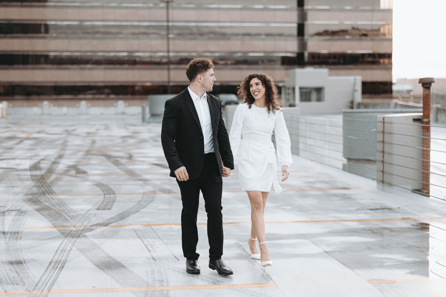A couple in formal attire walking together on an urban rooftop parking structure.