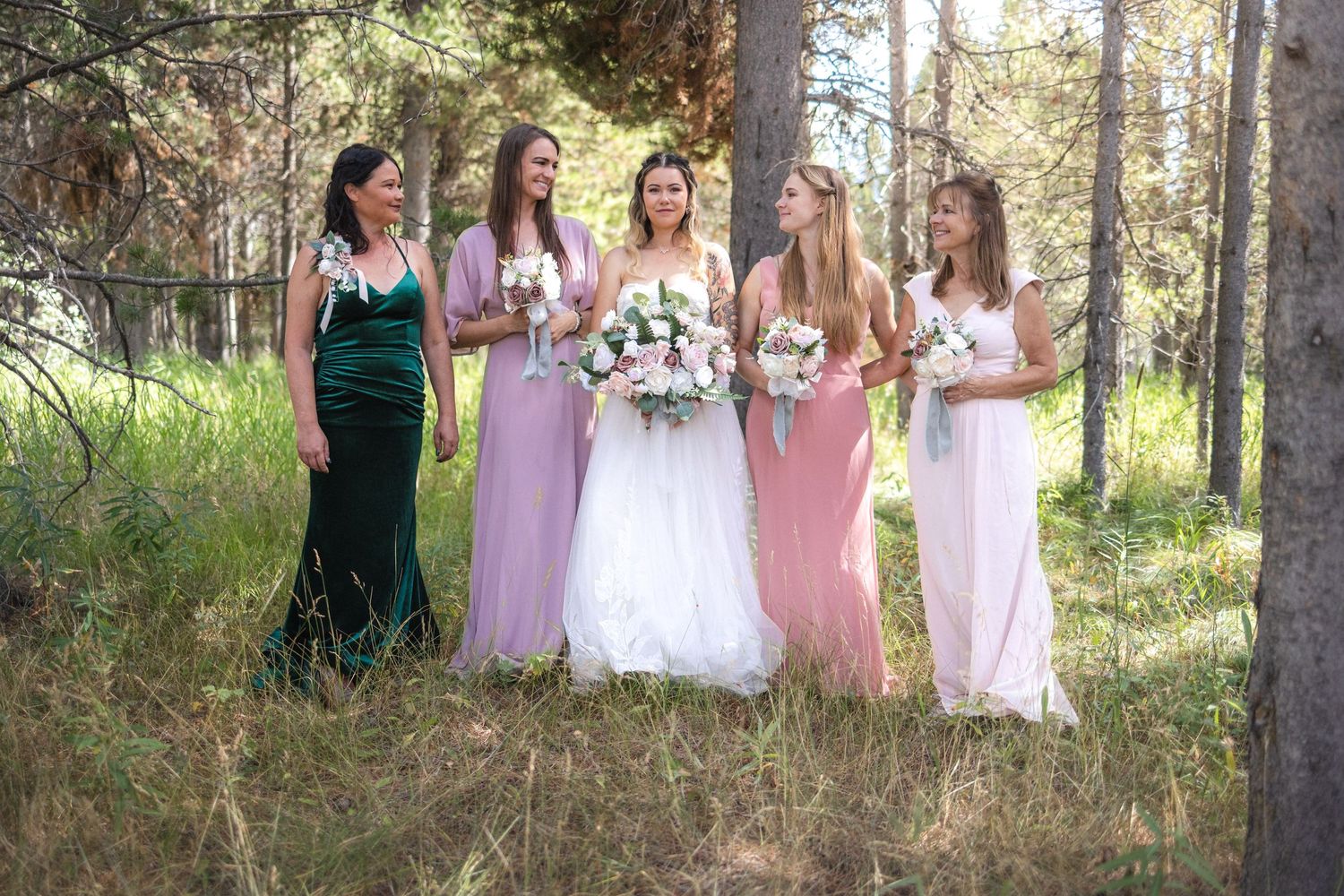 Wedding party lineup with bridesmaids in pink and green dresses holding floral bouquets in forest.