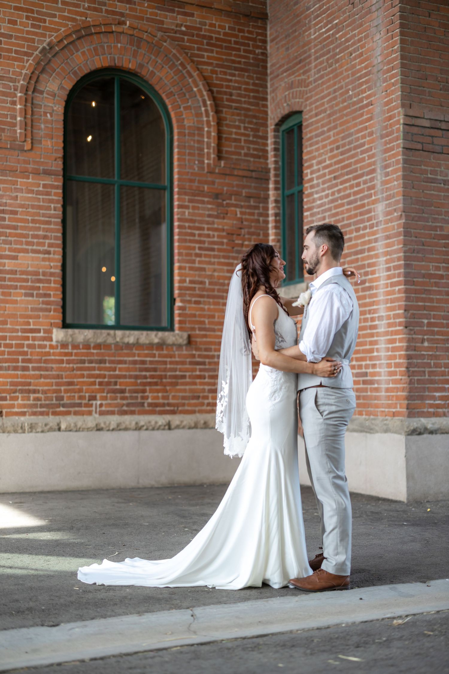 A romantic wedding couple embraces outside a historic brick building with arched windows.