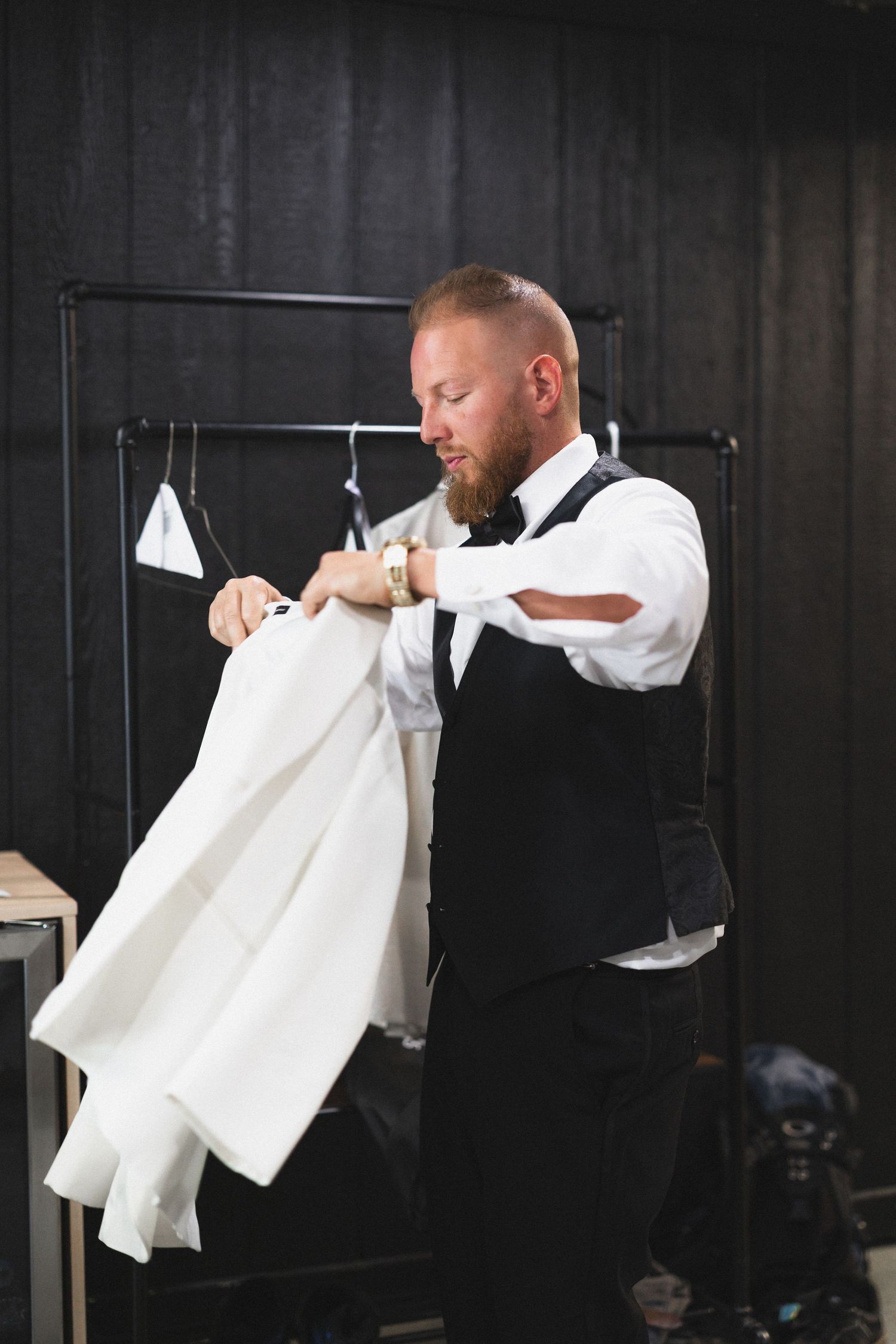 Groom in black vest handling white formal jacket near clothing rack.