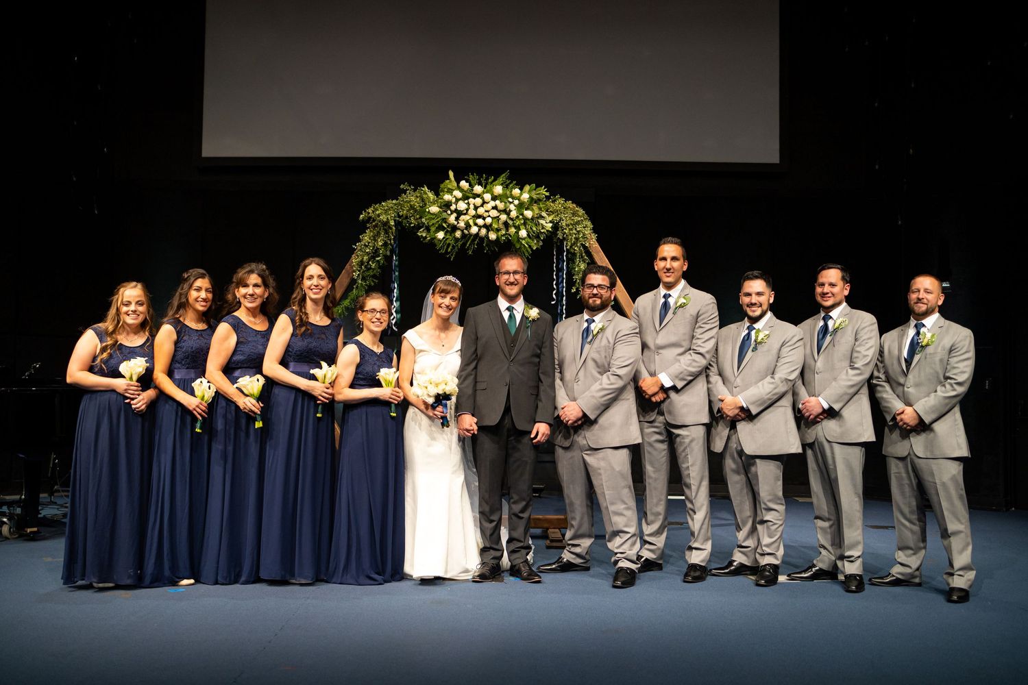 Wedding party poses for formal photo with bridesmaids in navy and groomsmen in gray suits.
