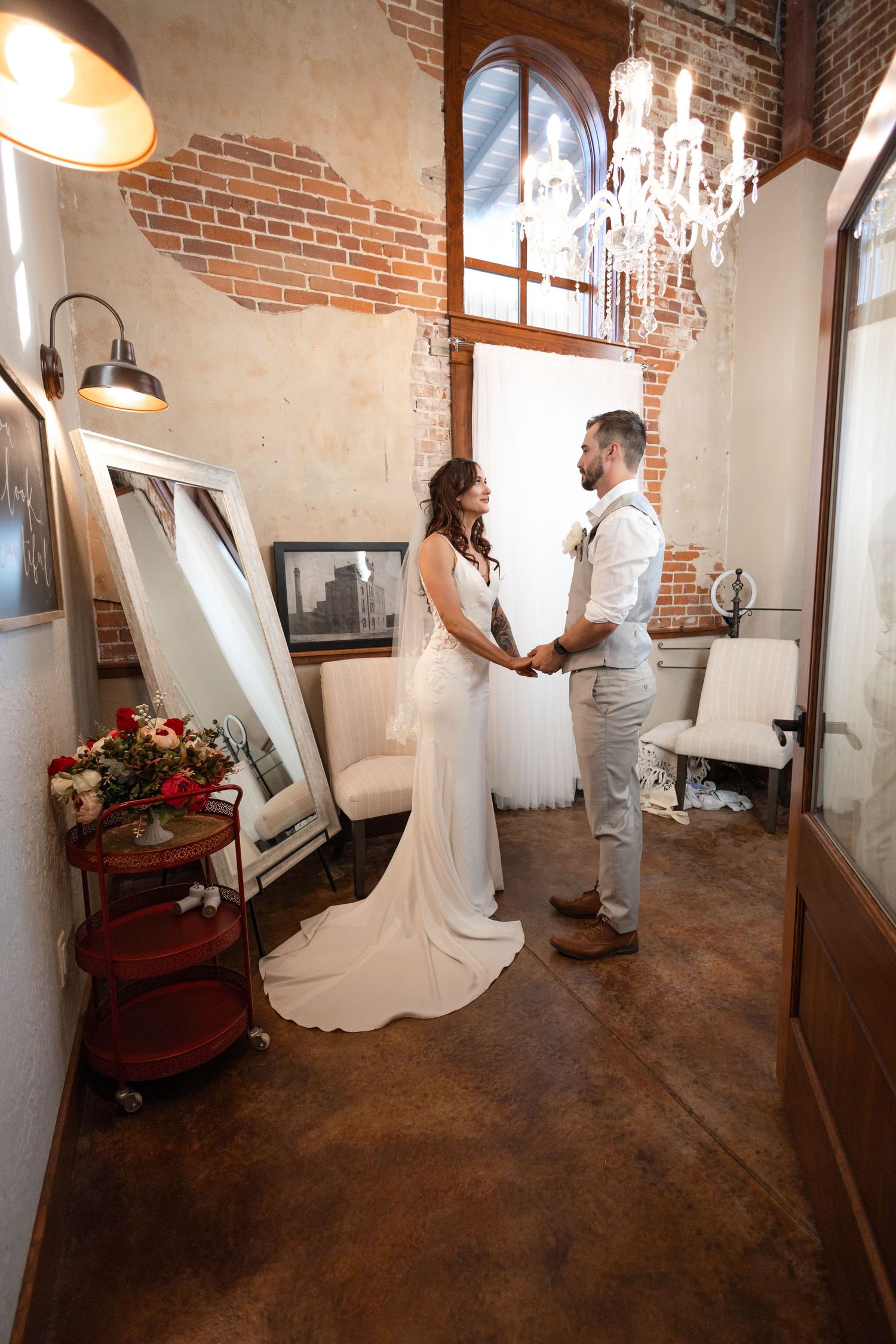 Wedding couple holds hands in vintage room with chandelier and exposed brick walls.