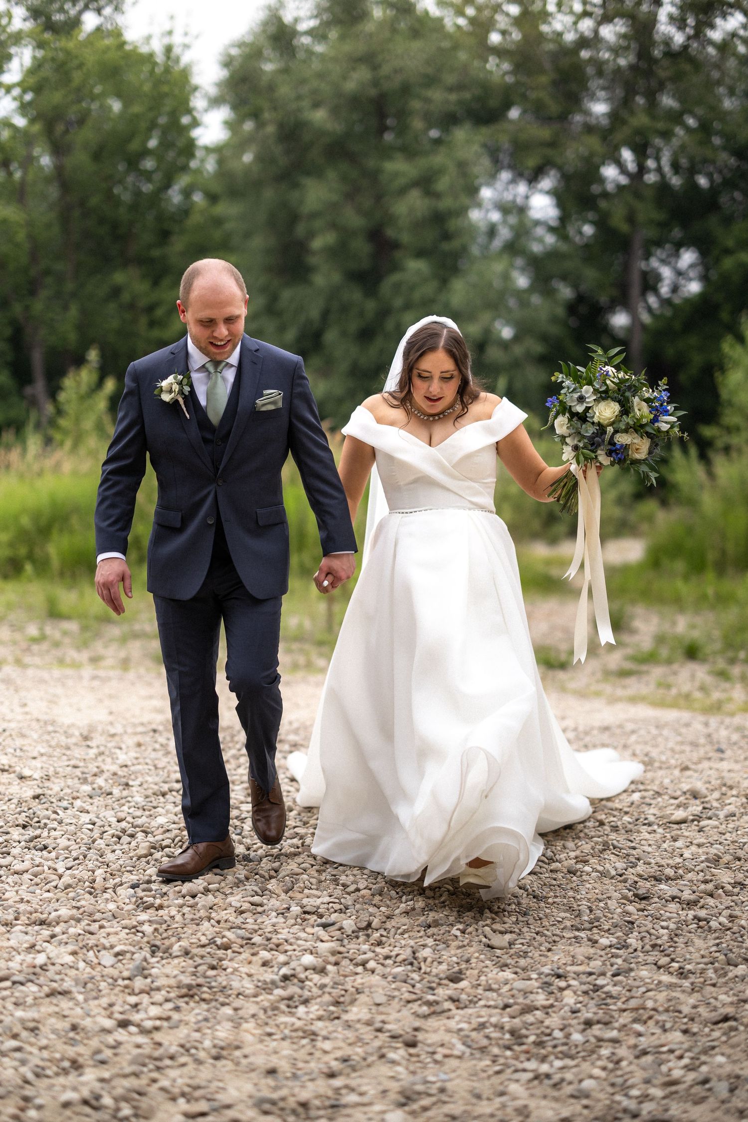 A couple walks together on a gravel path during their outdoor wedding, with the bride wearing an elegant white gown.