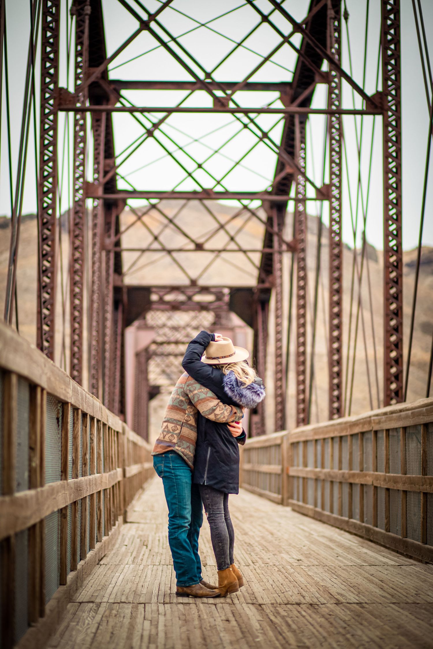 A couple embracing on a wooden bridge surrounded by metal trusses and beams.