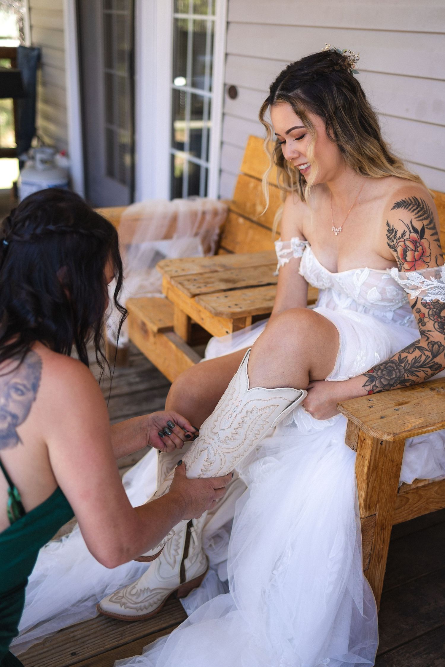 Bride with tattoos getting help putting on western-style boots before wedding ceremony.