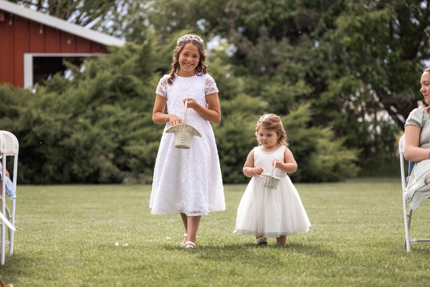 Two attendants in white dresses walk down a grassy aisle at an outdoor wedding ceremony.