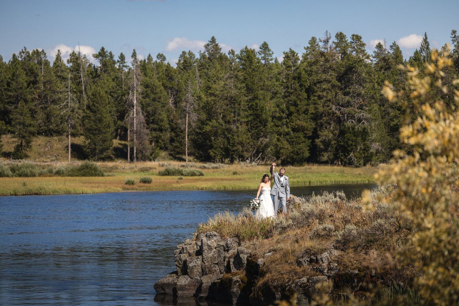 Wedding couple stands on rocky outcrop overlooking serene lake surrounded by pine forest.
