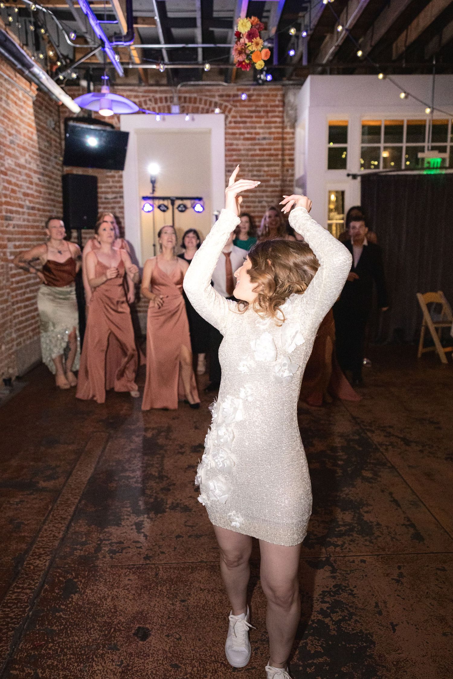 Energetic dancing and celebration at a wedding reception in a brick-walled venue with string lights.