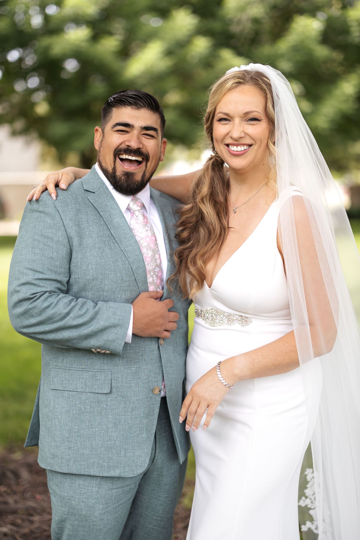 A couple in wedding attire poses together outdoors, with bride in white gown and groom in gray suit.