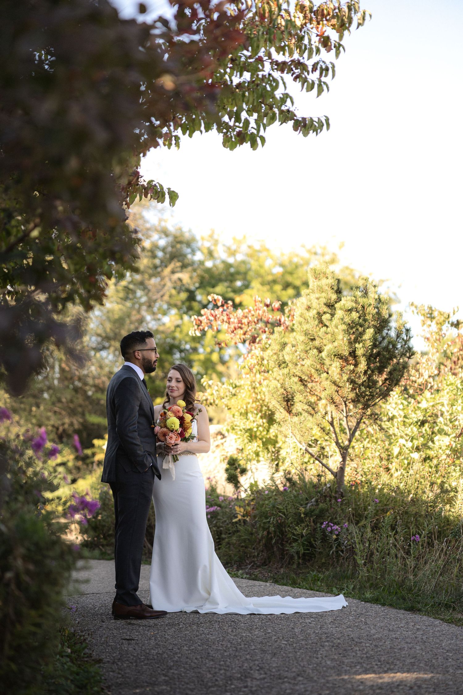 Full-length wedding portrait of couple standing on garden path in golden afternoon light.
