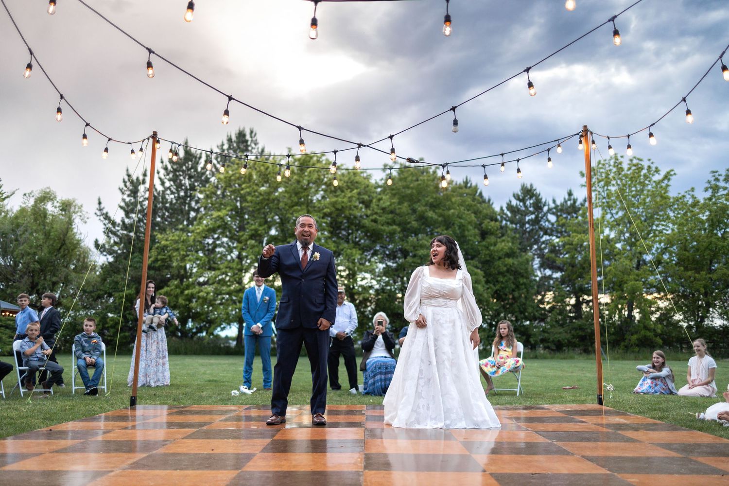 Wedding couple dancing on a wooden outdoor dance floor with string lights overhead.