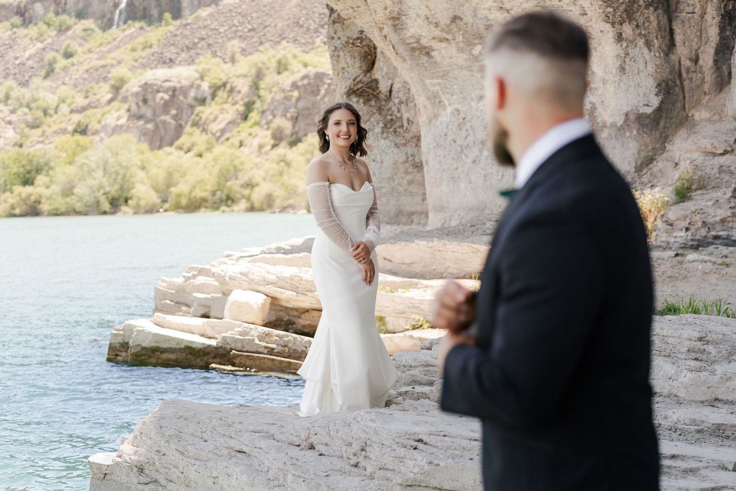 Bride in white lace gown smiles at groom in navy suit during outdoor wedding ceremony by a rocky riverside.
