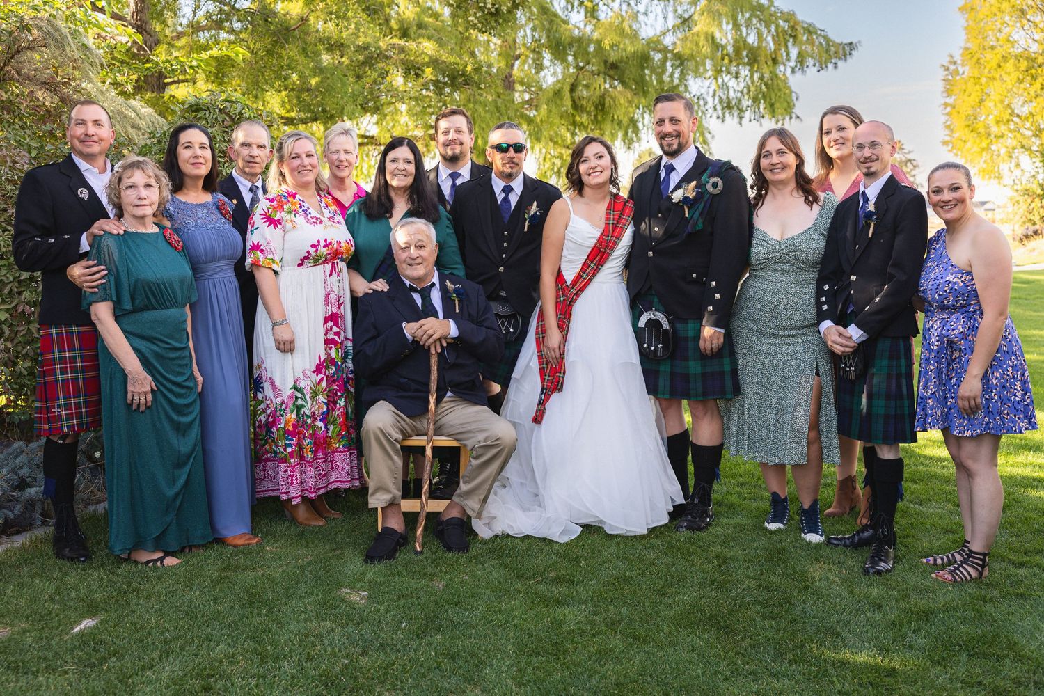 Large wedding party group poses outdoors with mix of kilts, formal wear and colorful dresses.
