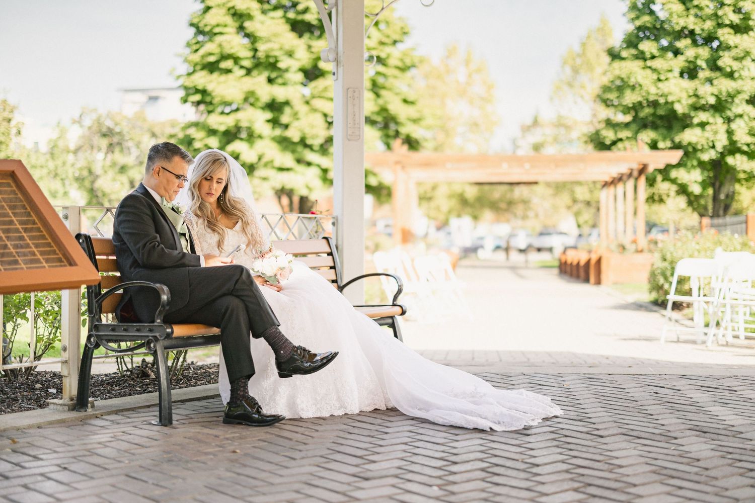 A couple sits together on a park bench at a wedding venue with landscaped grounds visible in the background.