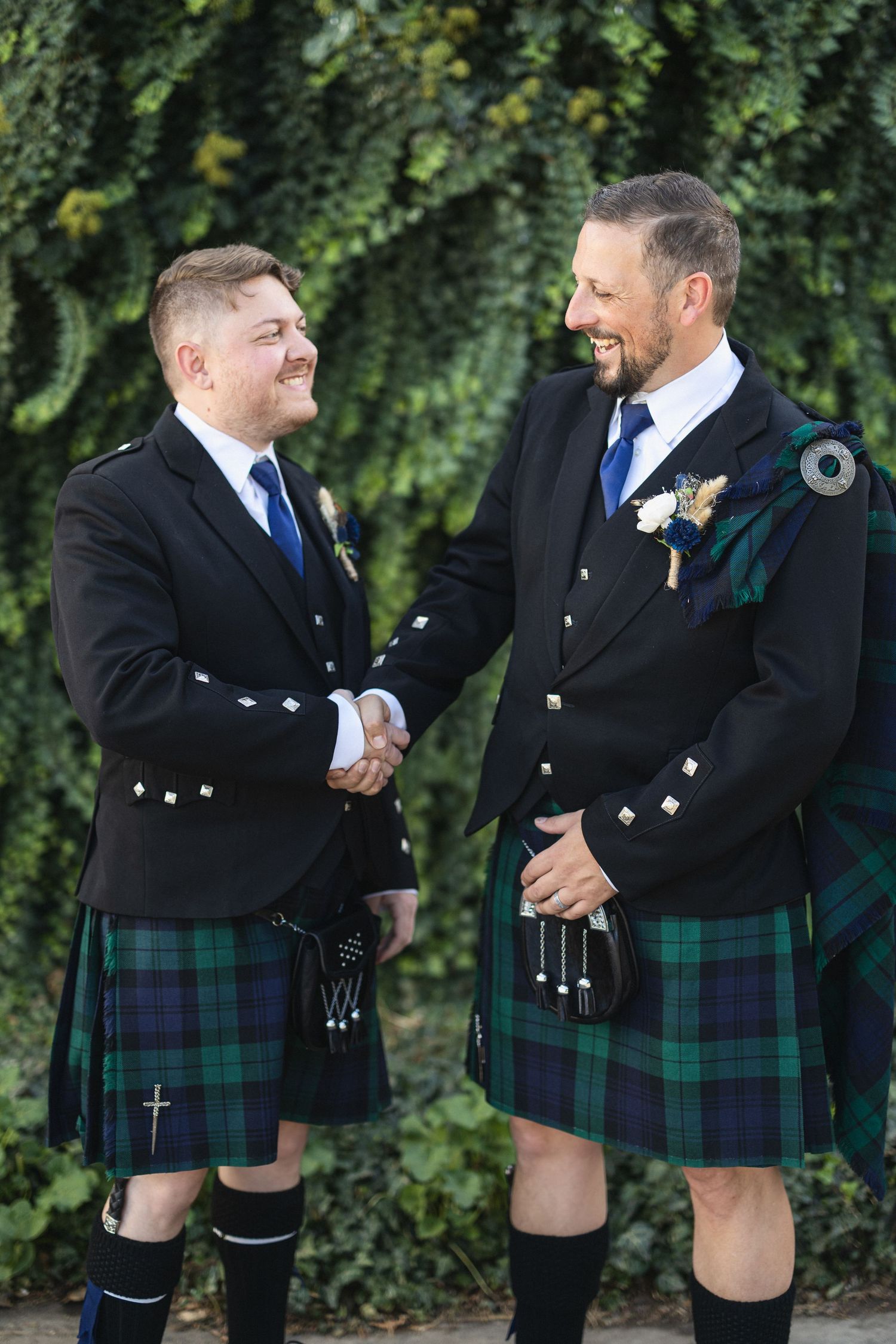 Two men in traditional Scottish kilts share handshake and laugh against green foliage.