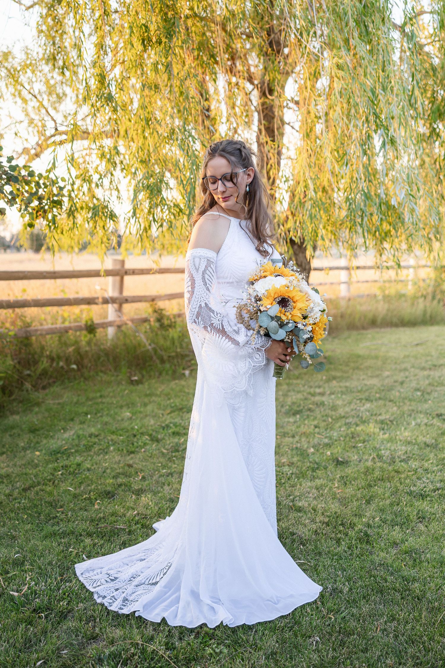 Bride in white lace dress holding sunflower bouquet poses under weeping willow trees at sunset.