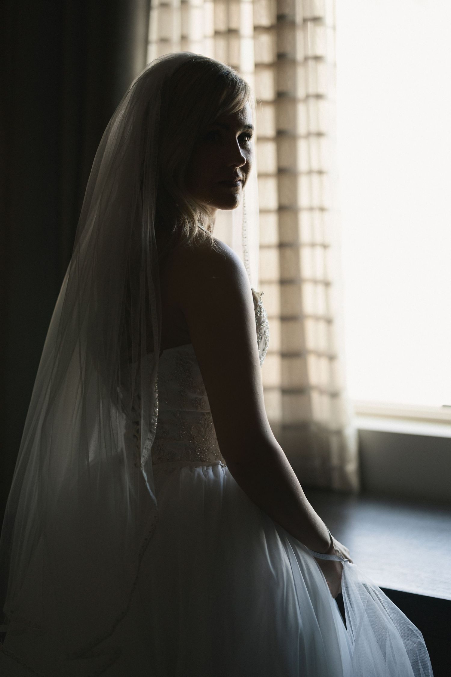 Dramatic silhouette of bride in wedding gown against sunlit window.