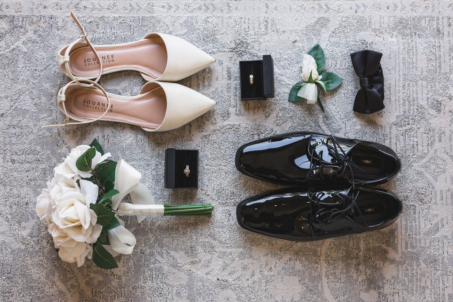 Flat lay of bride and groom shoes with ring boxes and white roses.