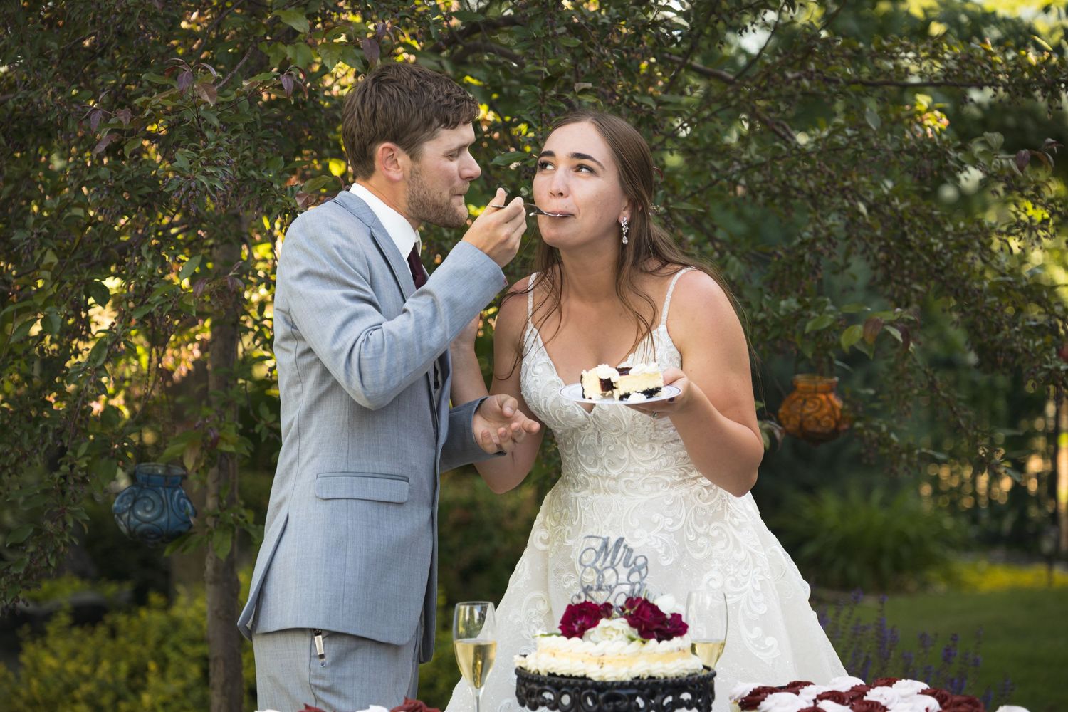 A couple shares wedding cake during their outdoor reception under string lights.