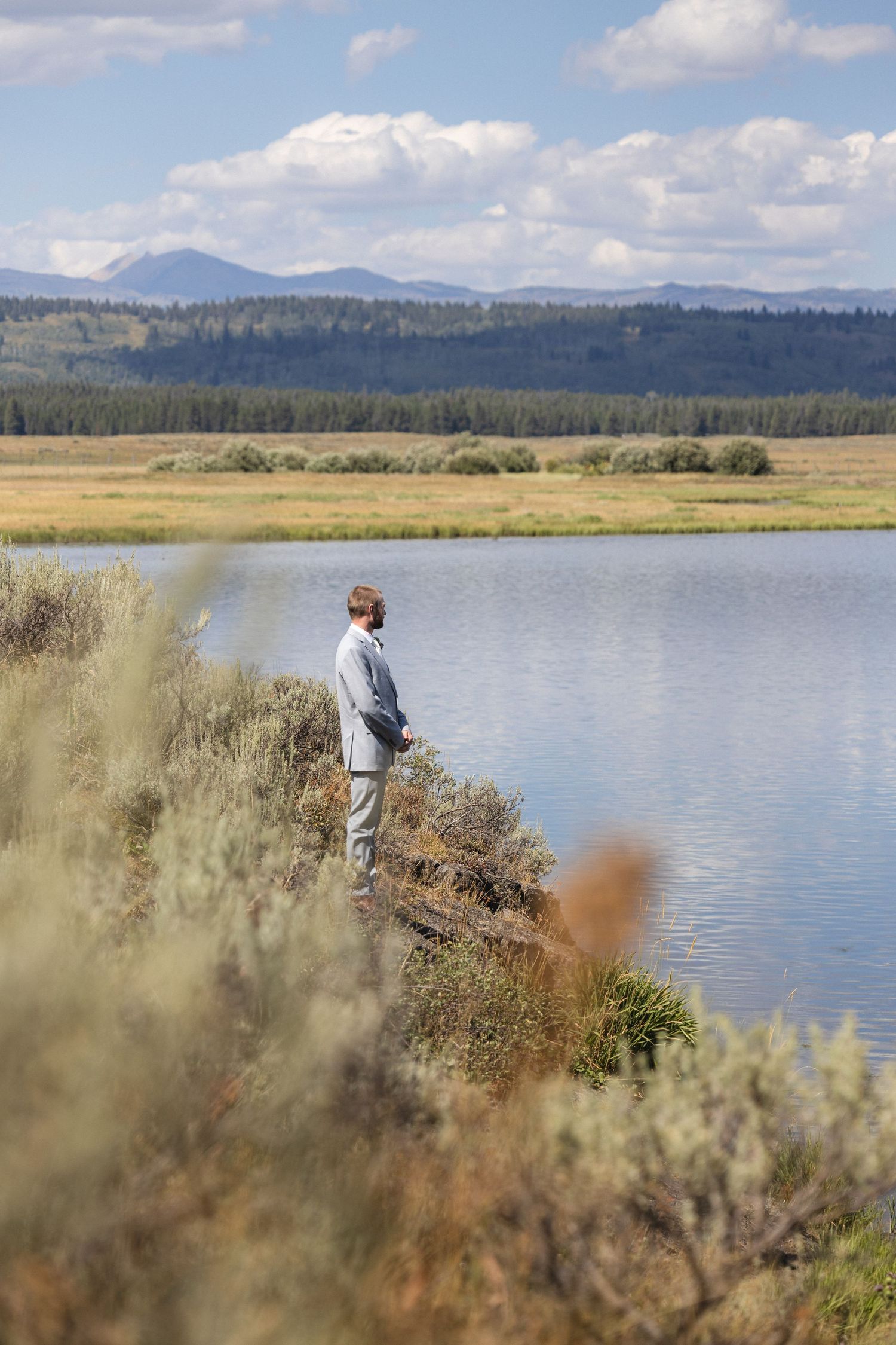 Person standing alone on lakeside cliff with mountains and forest in the background.