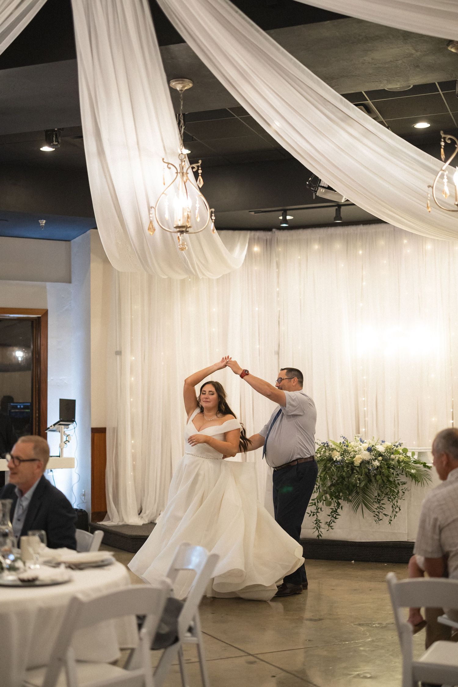 Couple dancing under draped white fabric and chandeliers at wedding reception.